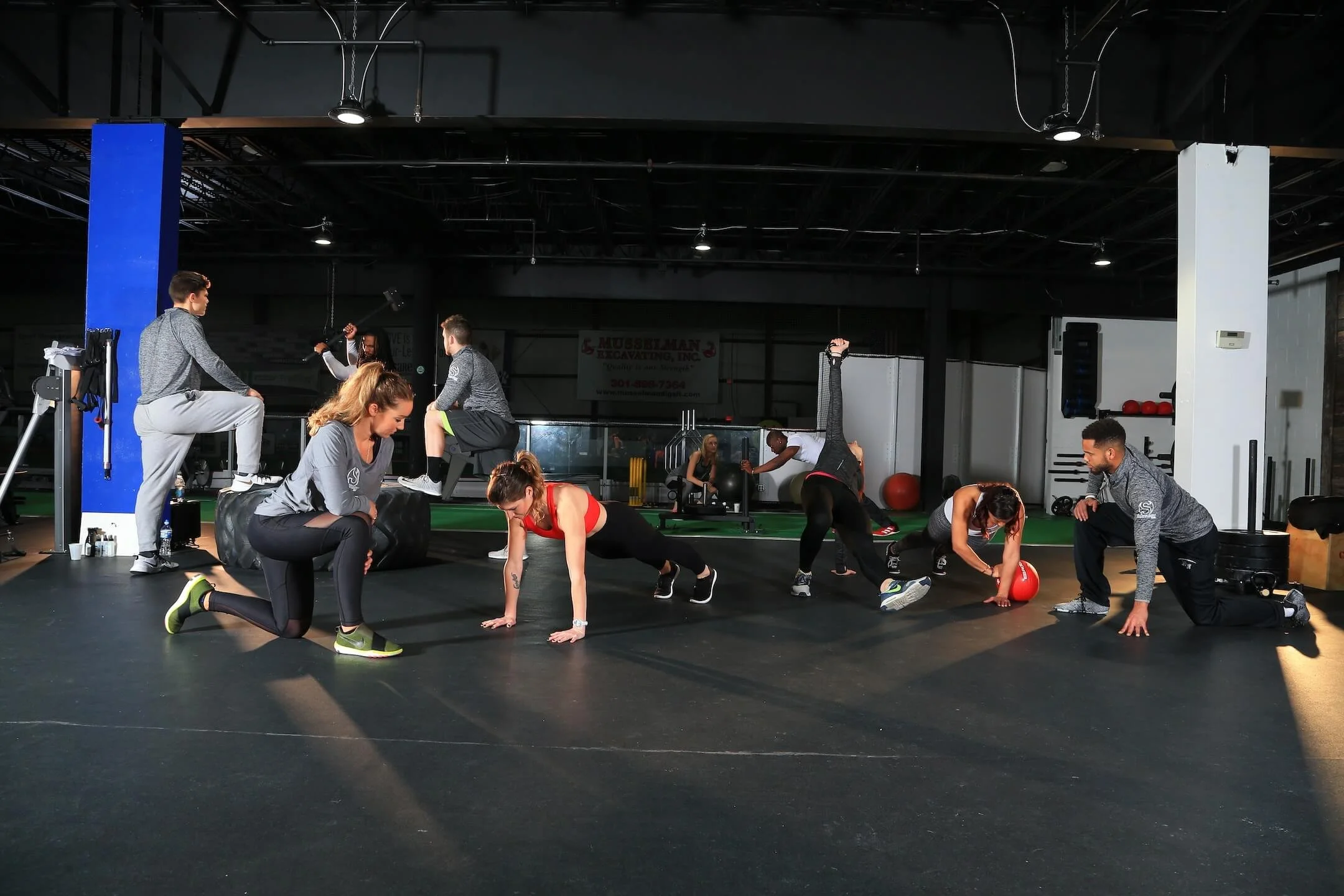 People exercising in a gym, performing various workouts on a black floor, including lunges, planks, and stretches, with gym equipment in the background.