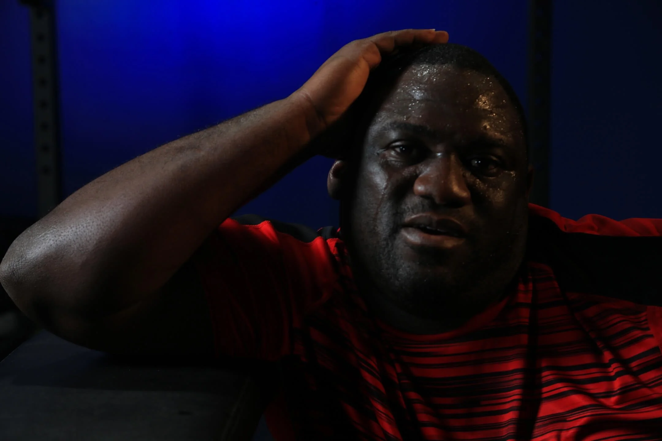 A man with dark skin, sweating, wearing a red and black striped sports shirt, resting his hand on his head in a dimly lit environment with a blue background.