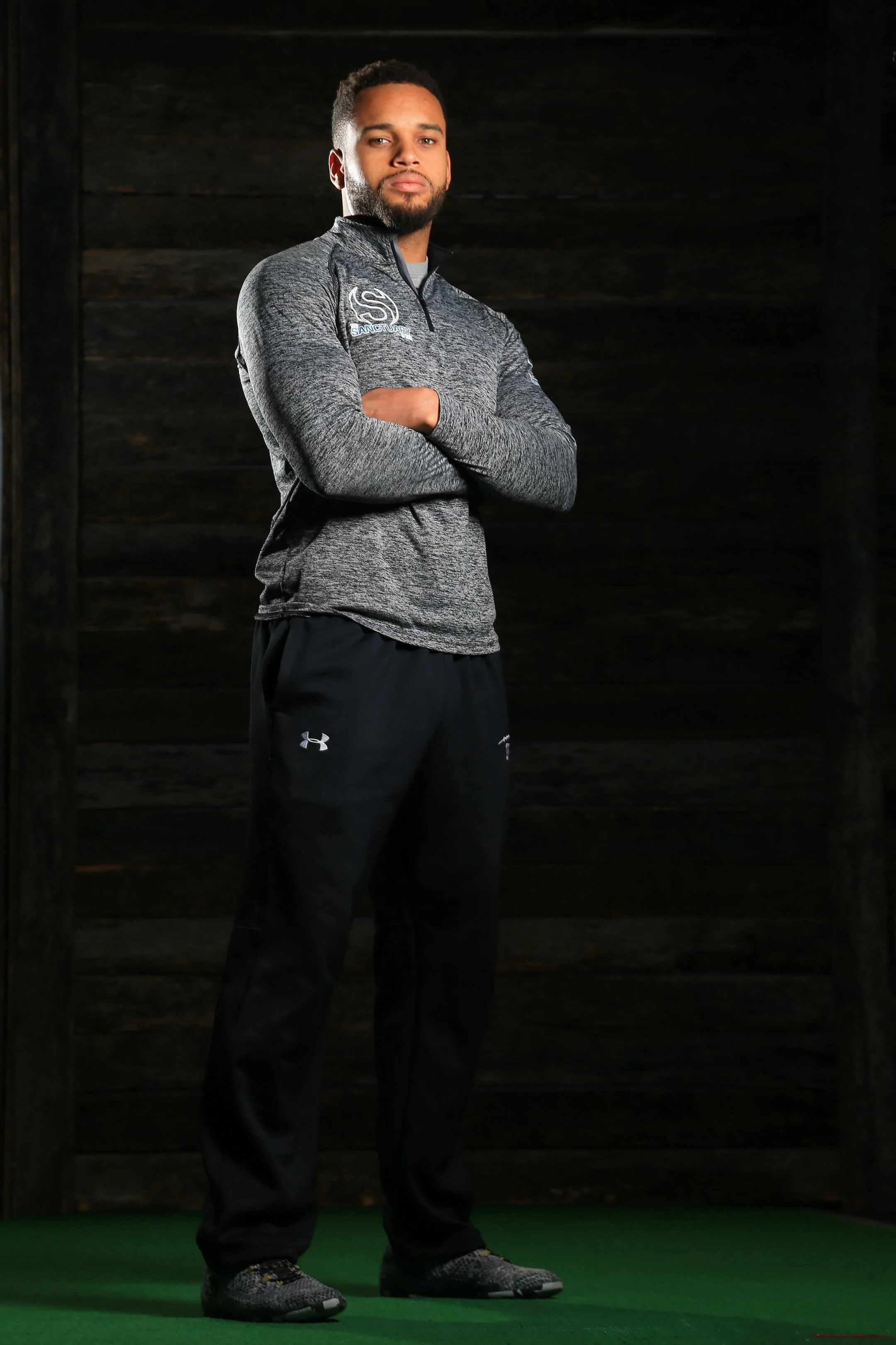 A man standing with arms crossed, wearing a gray zip-up athletic jacket, black Under Armour pants, and sneakers, in front of a dark wooden background.
