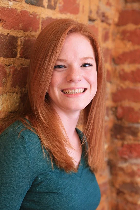 Headshot woman with red hair against brick wall wearing green sweater