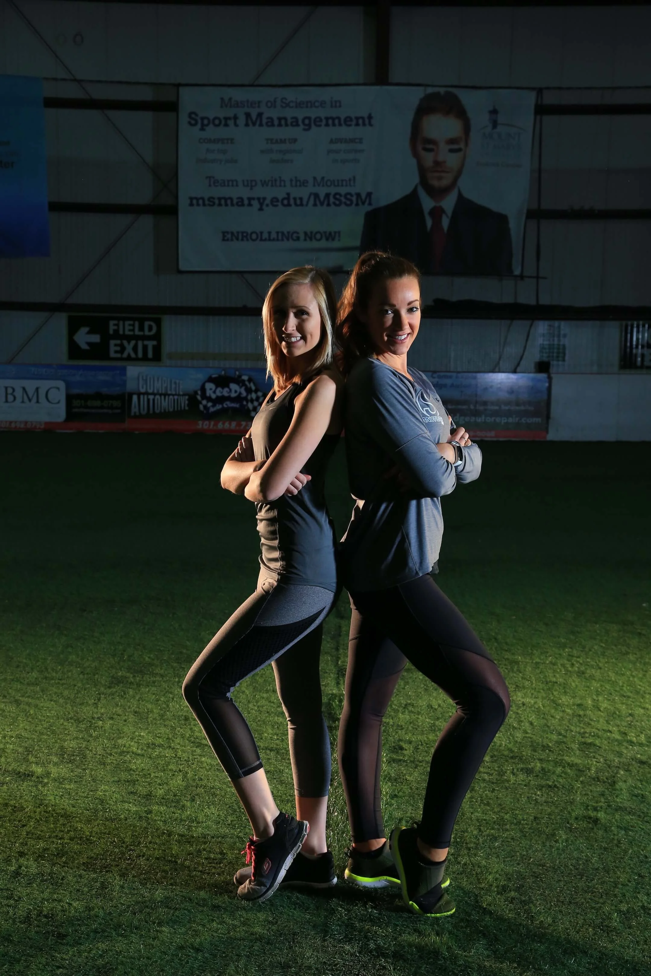 Two young women in athletic clothing standing back to back with arms crossed on a sports field, lit with artificial lighting, with promotional banners and a scoreboard in the background.