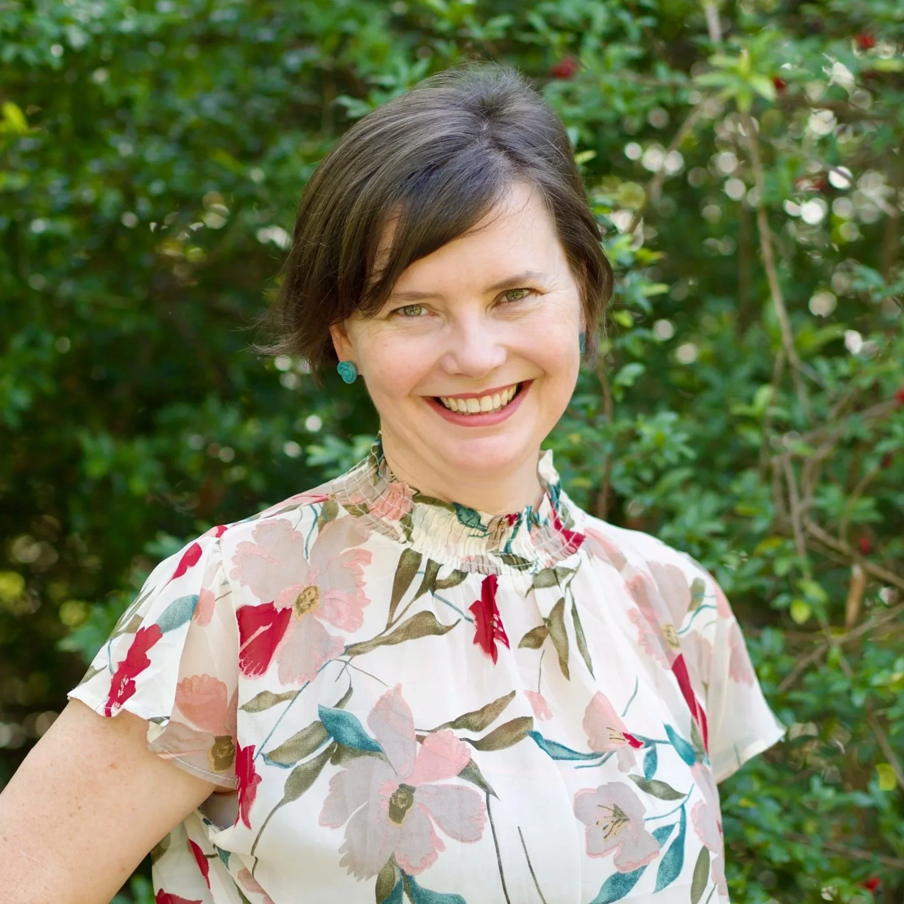 Brisbane counsellor, therapist. A woman smiling outdoors with green foliage in the background, wearing a floral blouse and turquoise earrings.