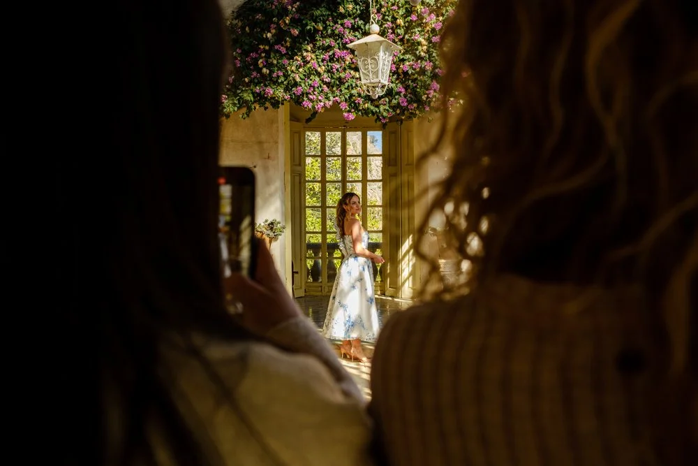 Mujer en vestido de novia en una habitación con ventanas y decoración floral, vista desde sombras de dos personas que la observan.