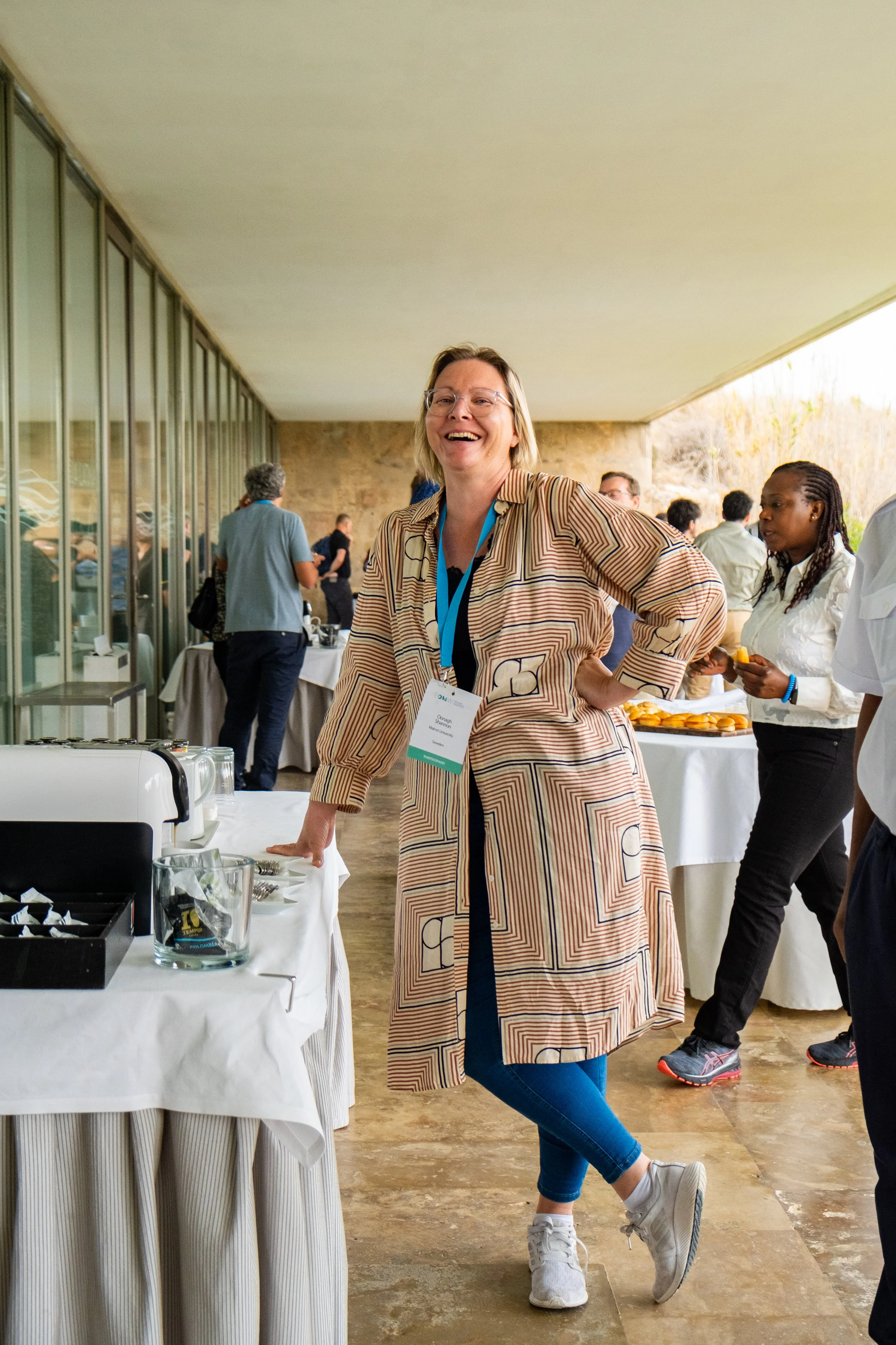 Mujer sonriendo en un evento social, con mesa de buffé y otras personas en el fondo.