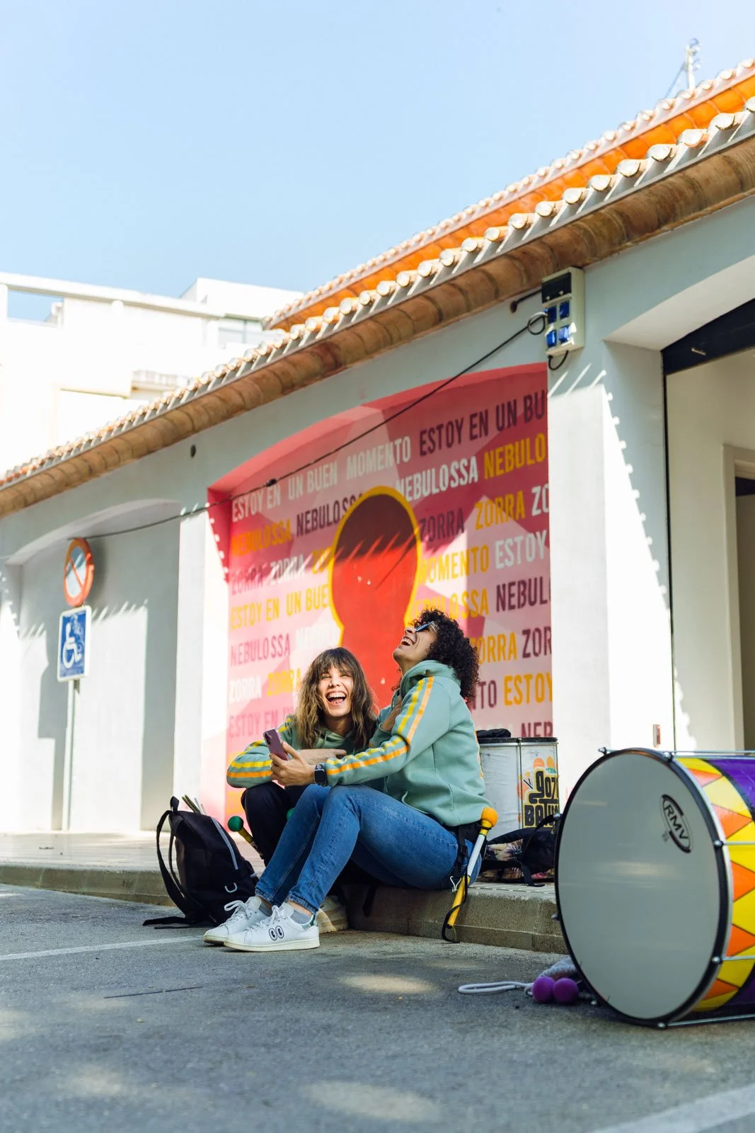 Dos mujeres sentadas en la acera riendo juntas, rodeadas de instrumentos musicales y equipo de banda, frente a un mural colorido en una pared.