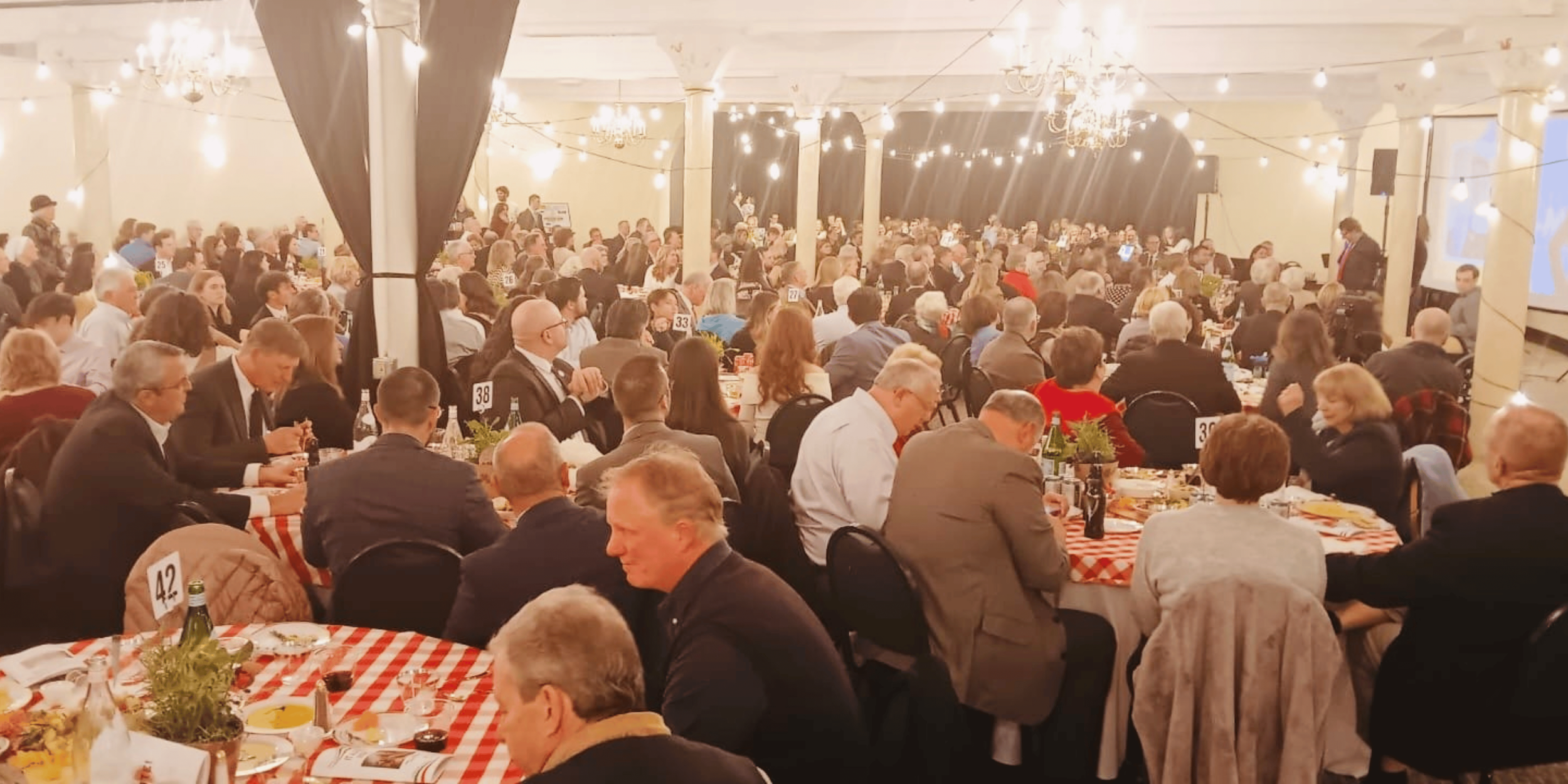 Large formal banquet with guests dining at numbered tables beneath chandeliers and string lights, with a stage and presentation screen.