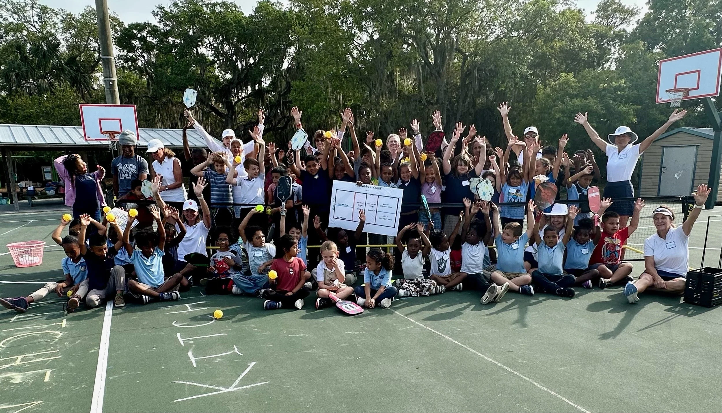 Large group of children and adults gathered on outdoor basketball court, some sitting and some standing, raising hands, holding tennis rackets and yellow tennis balls, with a basketball hoop in the background, trees, and a small building.