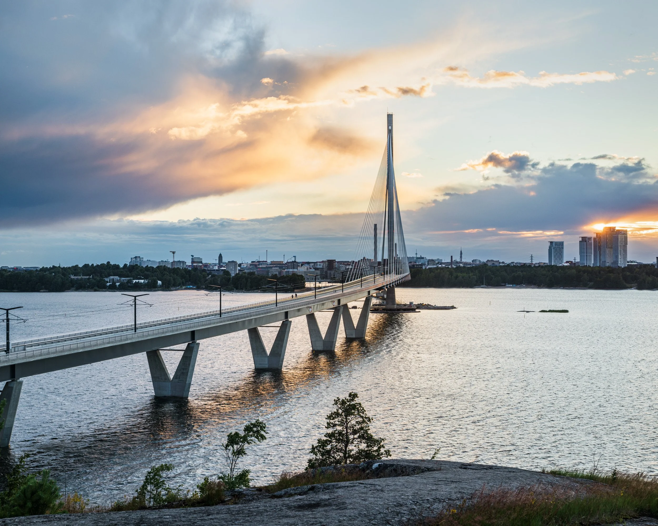 Helsingin Kruunuvuorensilta pilvisenä auringonlaskun hetkenä. Valo heijastuu mereen ja taustalla Helsingin kaupungin siluetti.