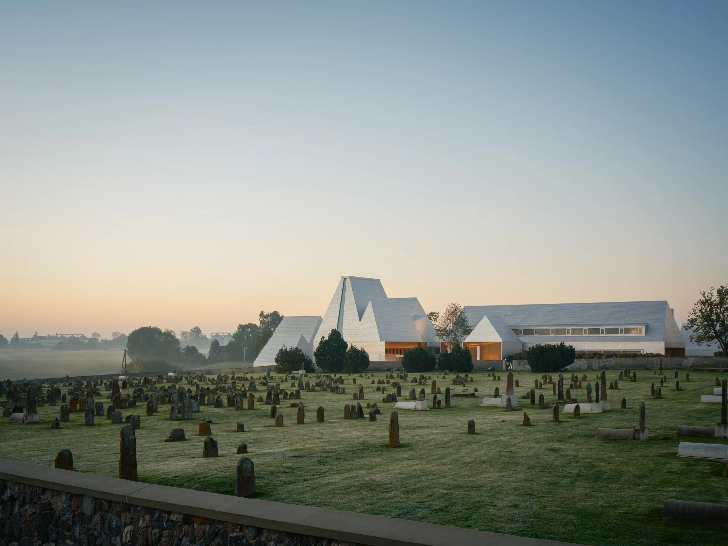 Moderni museorakennus maaseudulla nousee esiin vanhan hautausmaan keskeltä; vaaleasävyinen, kulmikas rakennus näkyy rauhallisessa maisemassa varhaisen aamun valossa.