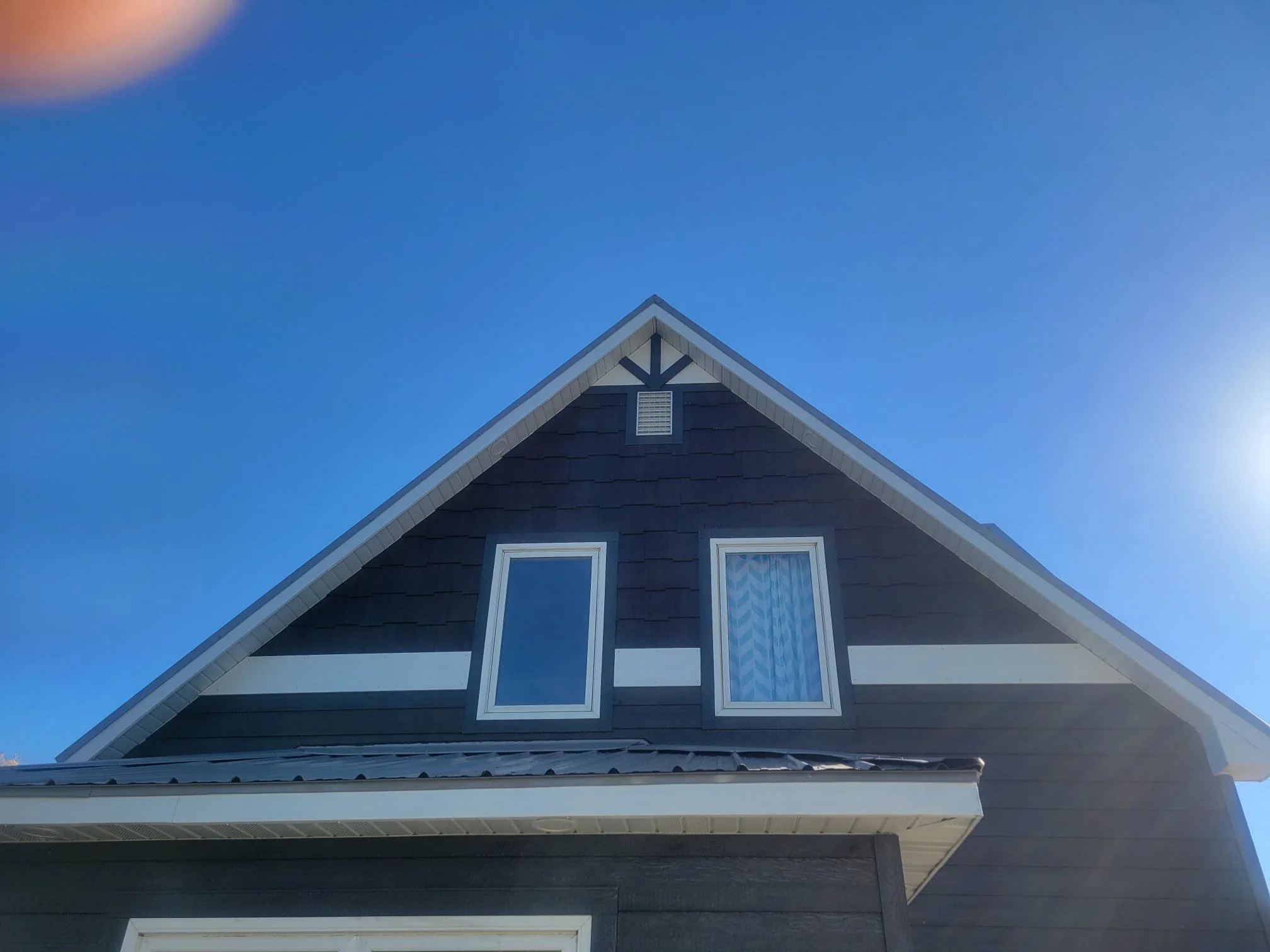 A close-up view of the upper part of a modern house with dark siding, white trim, two windows, and a steep pitched roof against a clear blue sky.