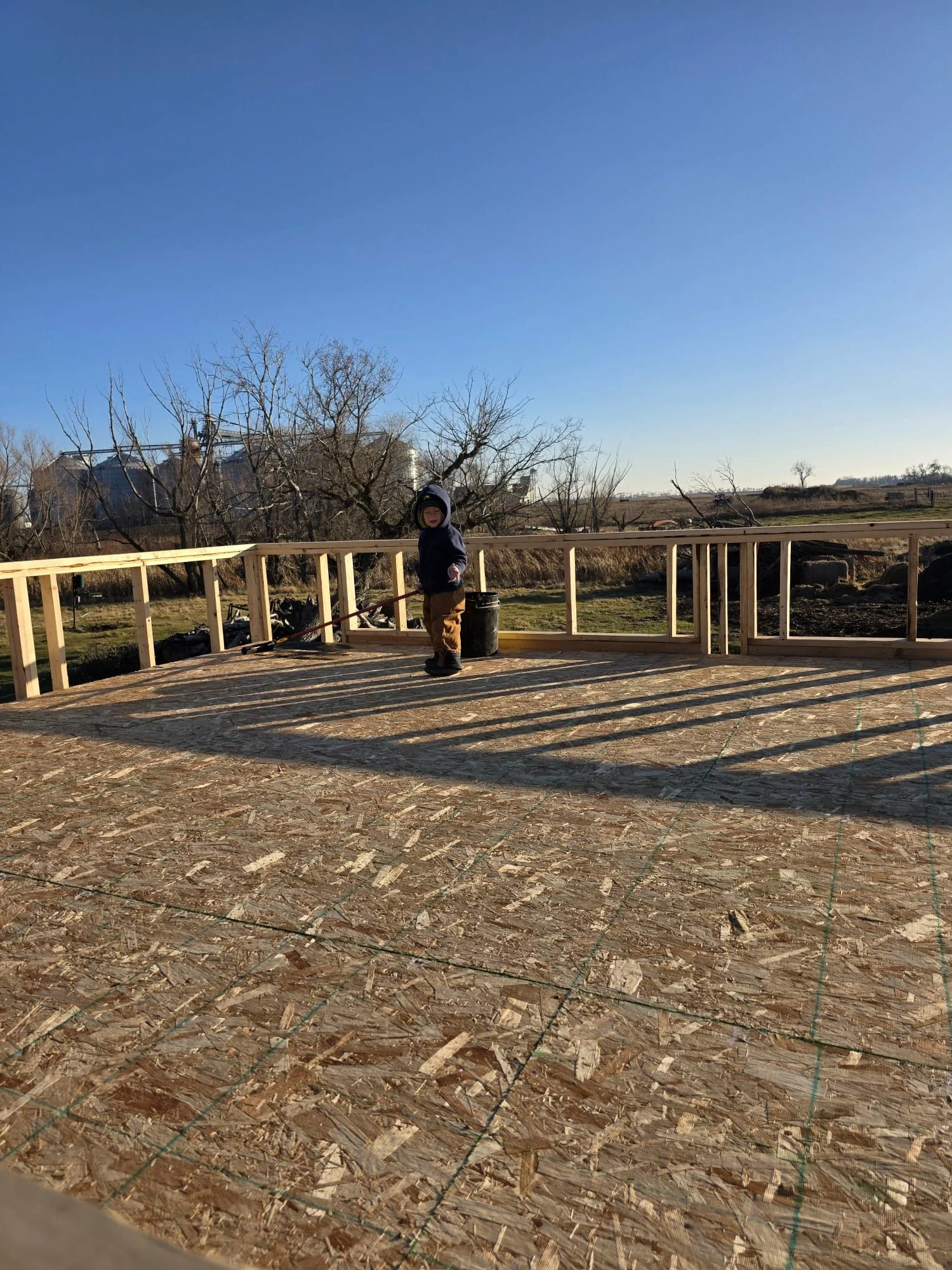 A young child dressed in a dark hoodie and tan pants standing on a construction site with unfinished wooden railing and plywood floor, holding a rake, with a clear blue sky and leafless trees in the background.