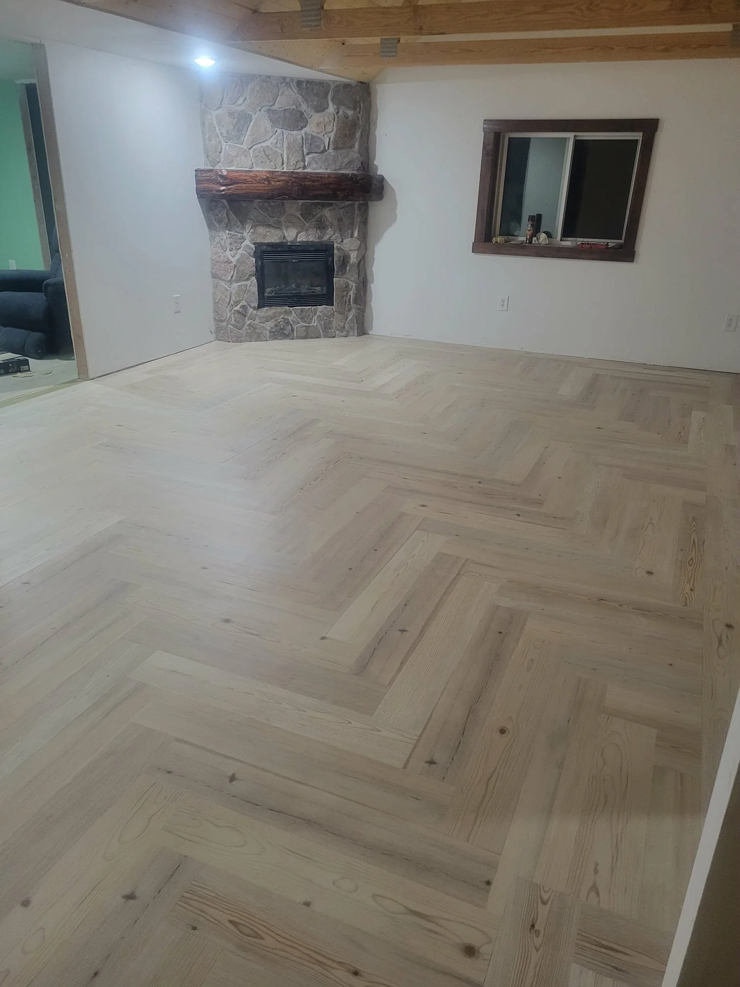 Empty living room with light-colored herringbone wood flooring, wood-beamed ceiling, stone fireplace with wooden mantle, and a window with a wooden frame on the wall.
