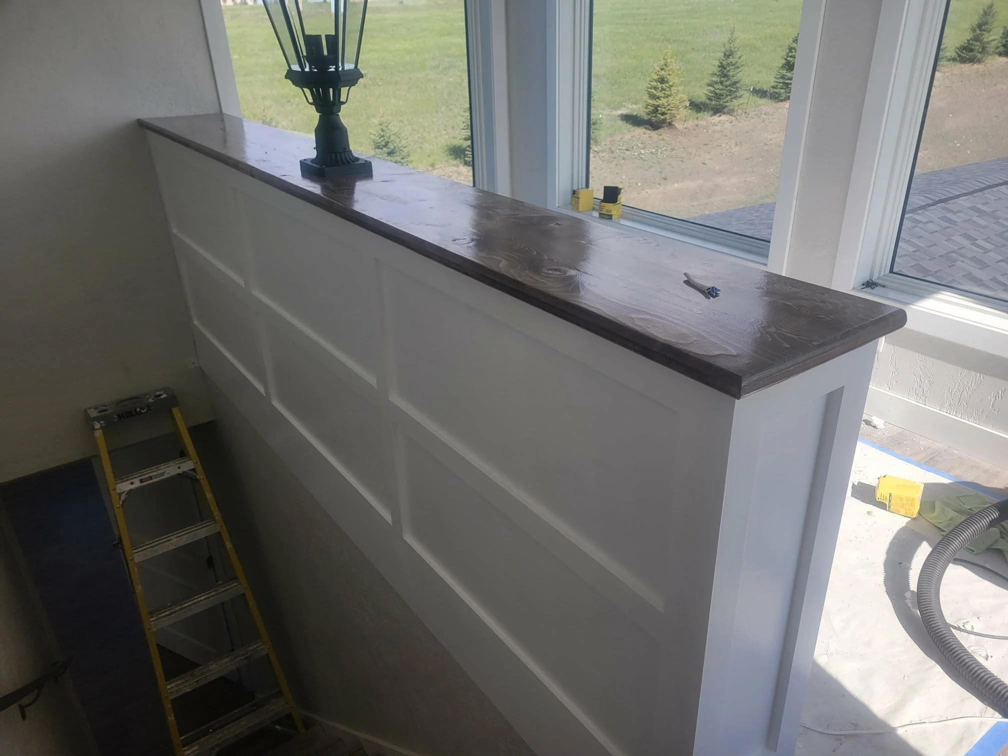 Interior view of a new built-in white cabinet or wall panel with a dark wood countertop, situated in front of large windows showing a backyard with trees and grass, a small ladder leaning against the wall, and construction tools and materials nearby.