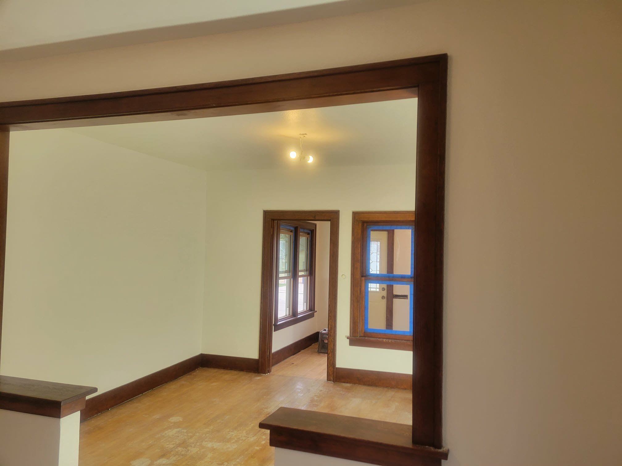 Empty room with wooden trim around windows and door, hardwood floor, yellowed walls, and a ceiling light fixture. 1920's house remodel.
