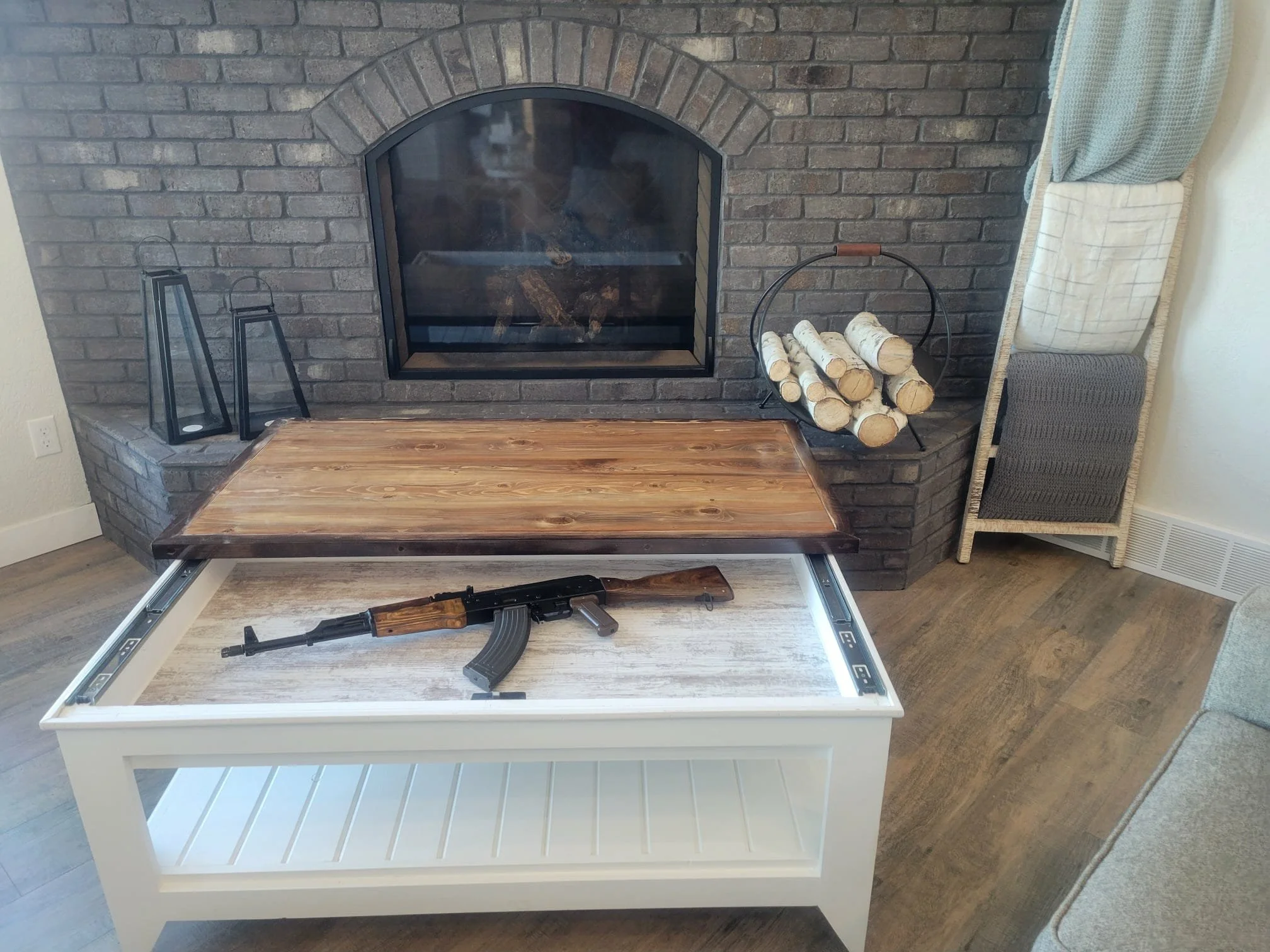 A room with a brick fireplace, a wooden table with a black rifle on top, and a white coffee table in front of it.