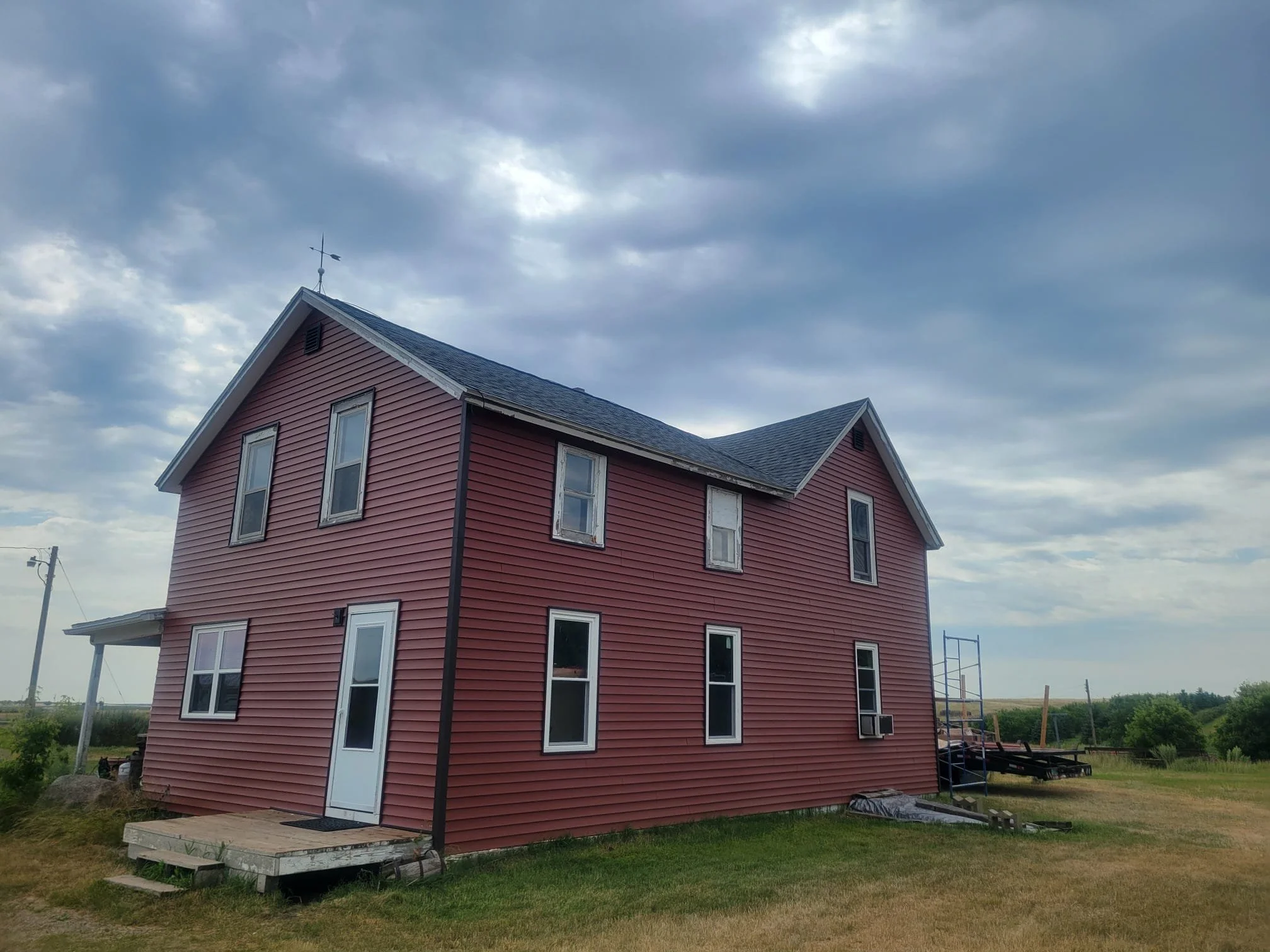 Red two-story house with multiple windows, a front door, and a small porch, set in a rural area under cloudy skies.