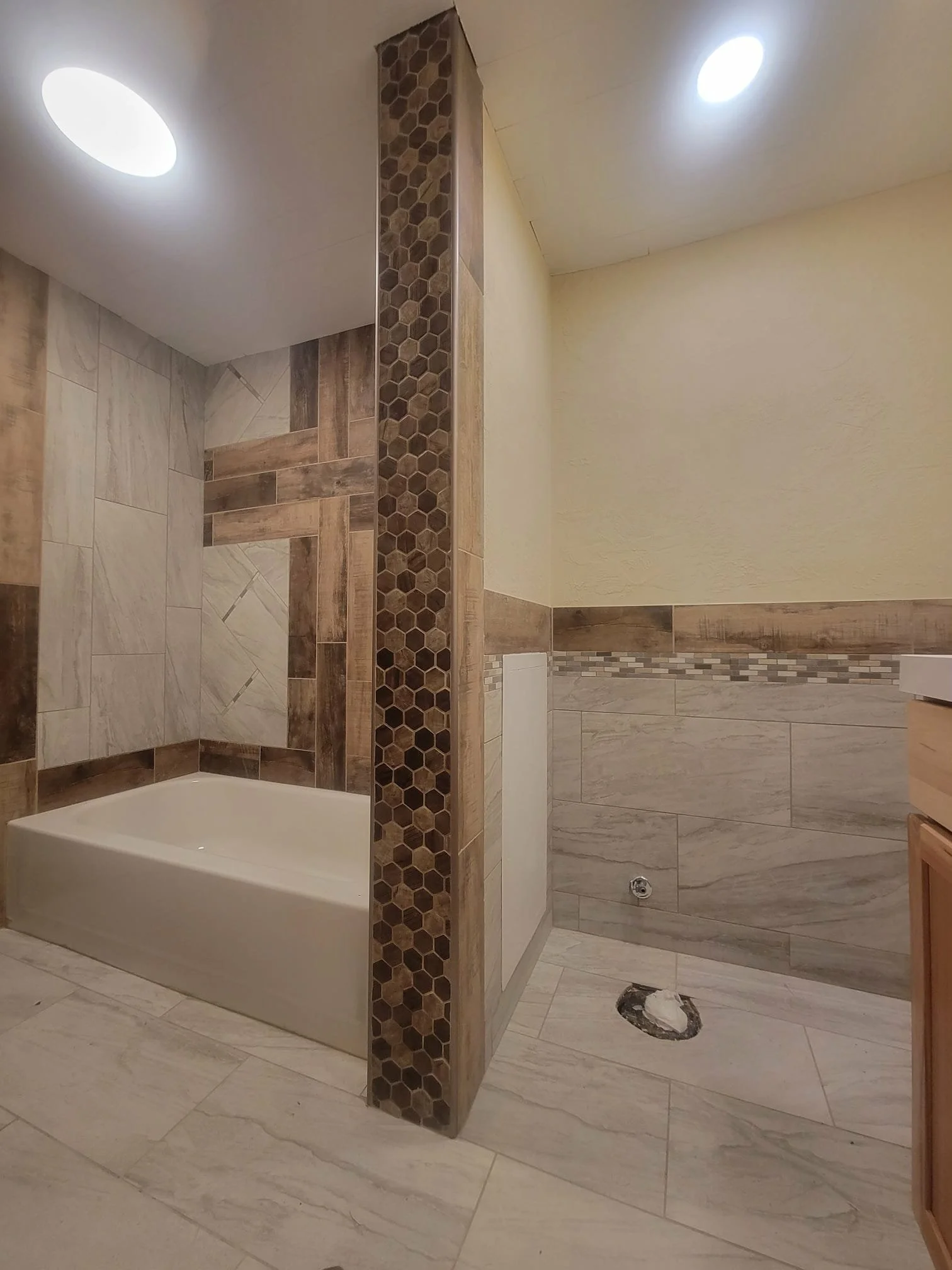 Bathroom under construction with a corner shower area and a separate wet area, tiled with beige and brown stone-like tiles, with visible plumbing fixtures and a partially finished wall.