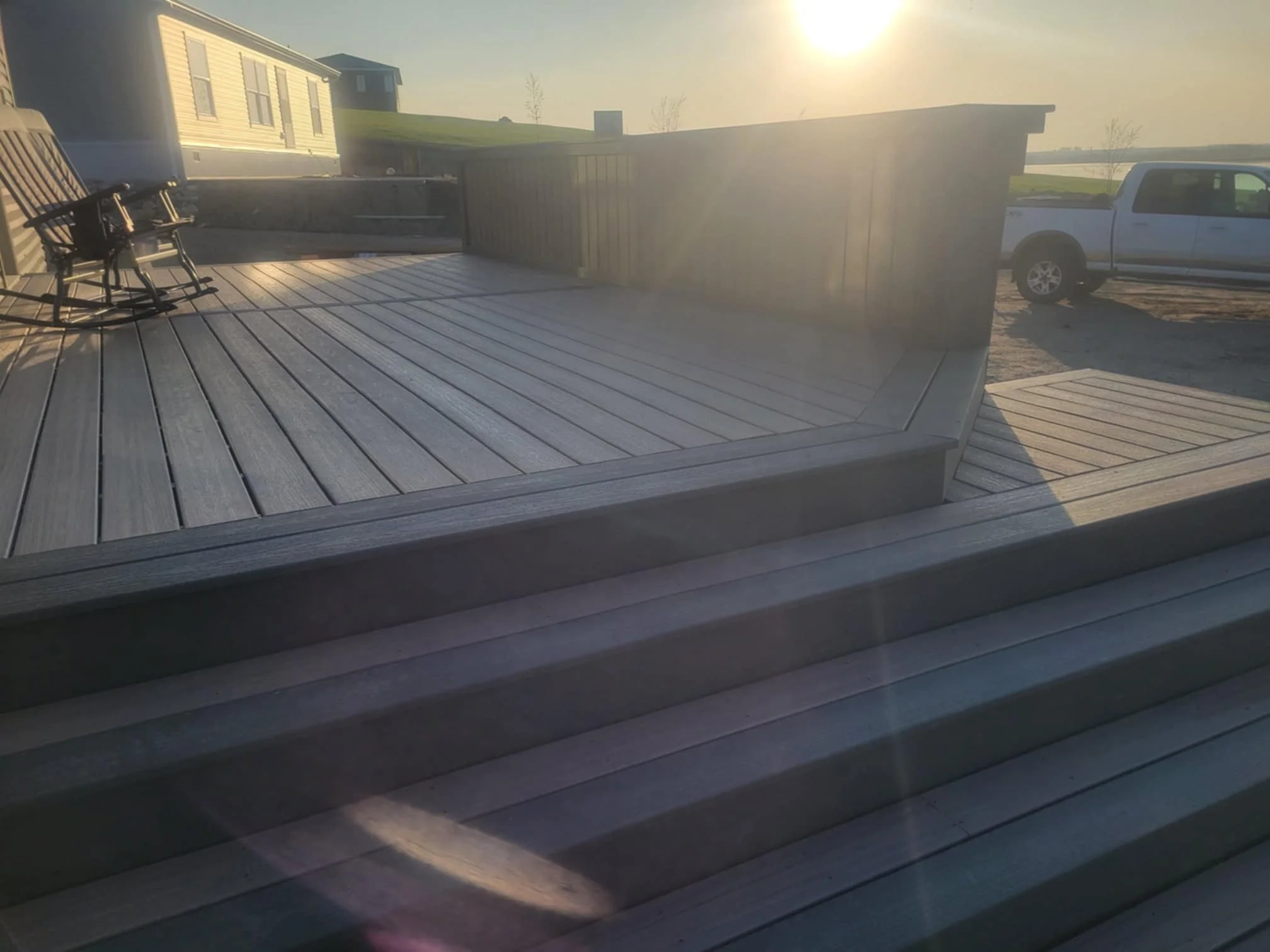 A view of a wooden deck with steps, a black metal bench with wooden slats, a wooden fence, and a white pickup truck parked on dirt. The background shows a grassy hill and a house, with the sun low in the sky.