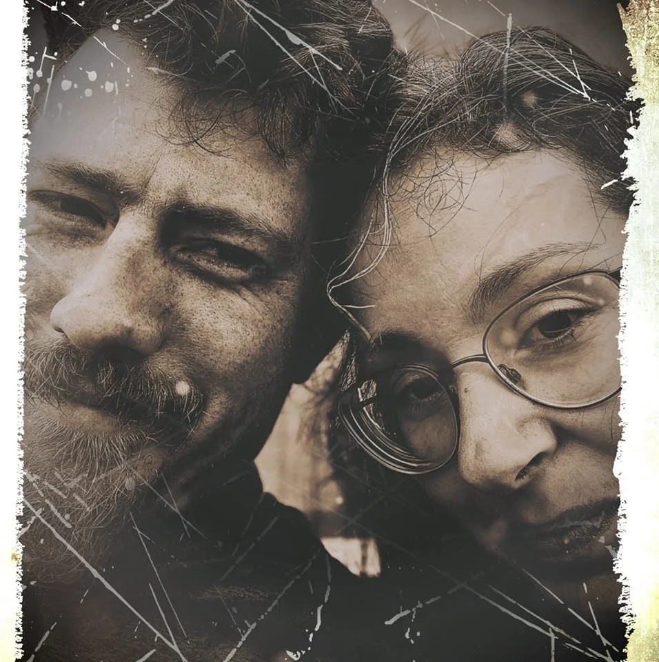 Close-up of a man and woman with rugged faces and glasses, both smiling and leaning toward each other, in a vintage-style photo with scratches and faded edges.