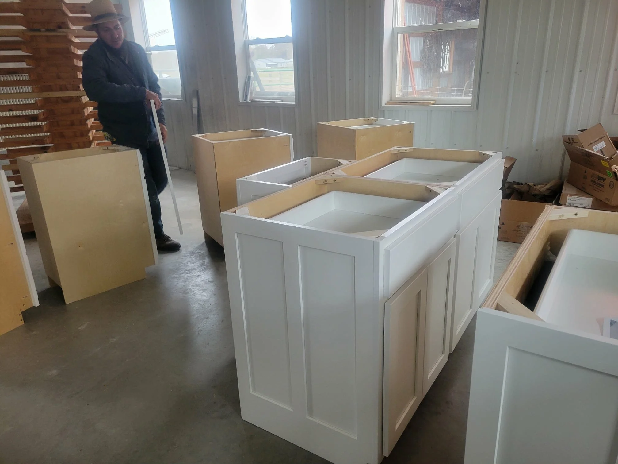 A person standing next to white kitchen cabinets inside a workshop, with several unassembled cabinets and boxes around.