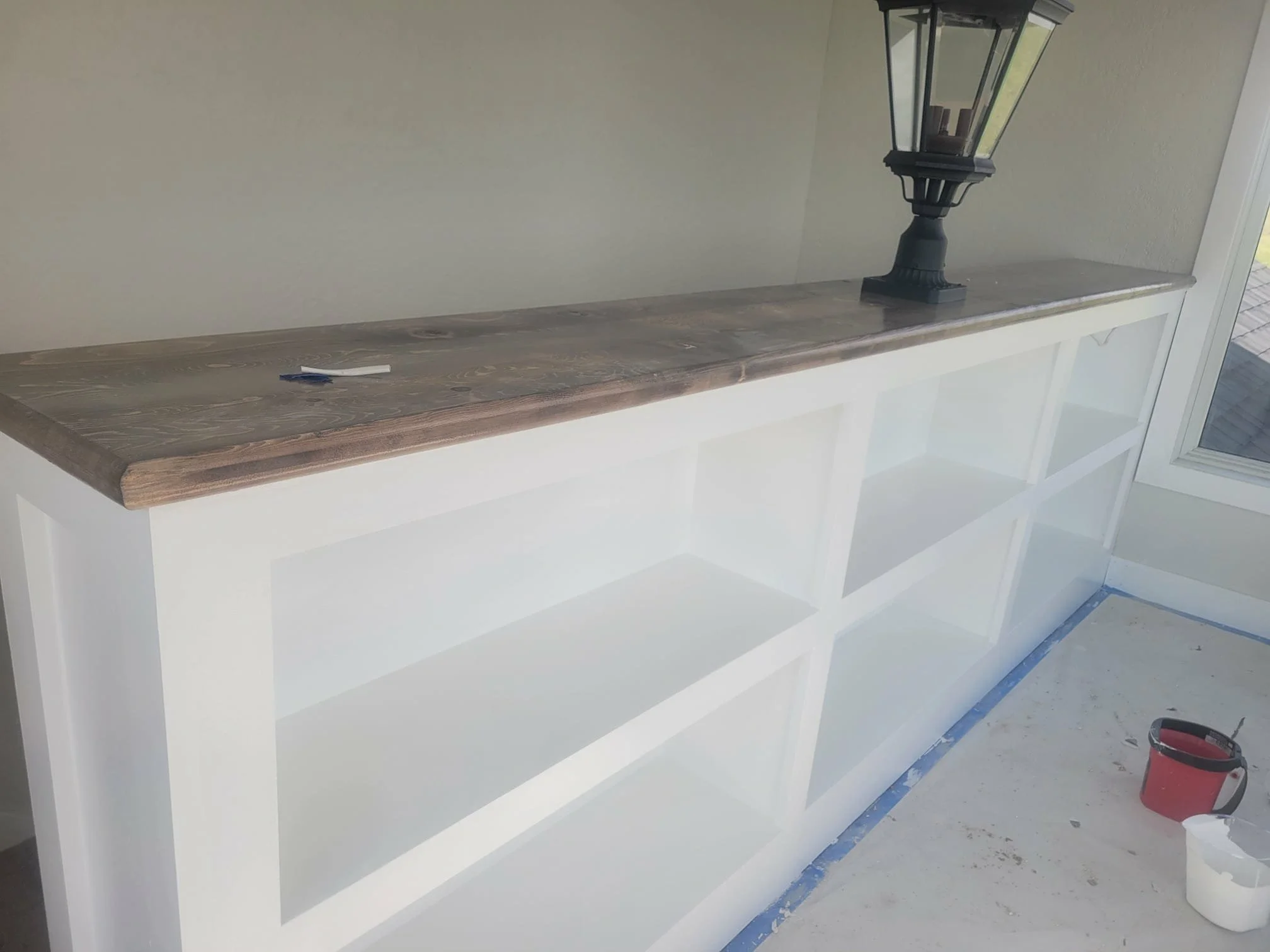 A wooden countertop on a white built-in shelf, with a black outdoor lantern-style lamp and a small color swatch on it. The area appears to be under construction or renovation, with a paint bucket and supplies on the floor and a sliding glass door to the right.
