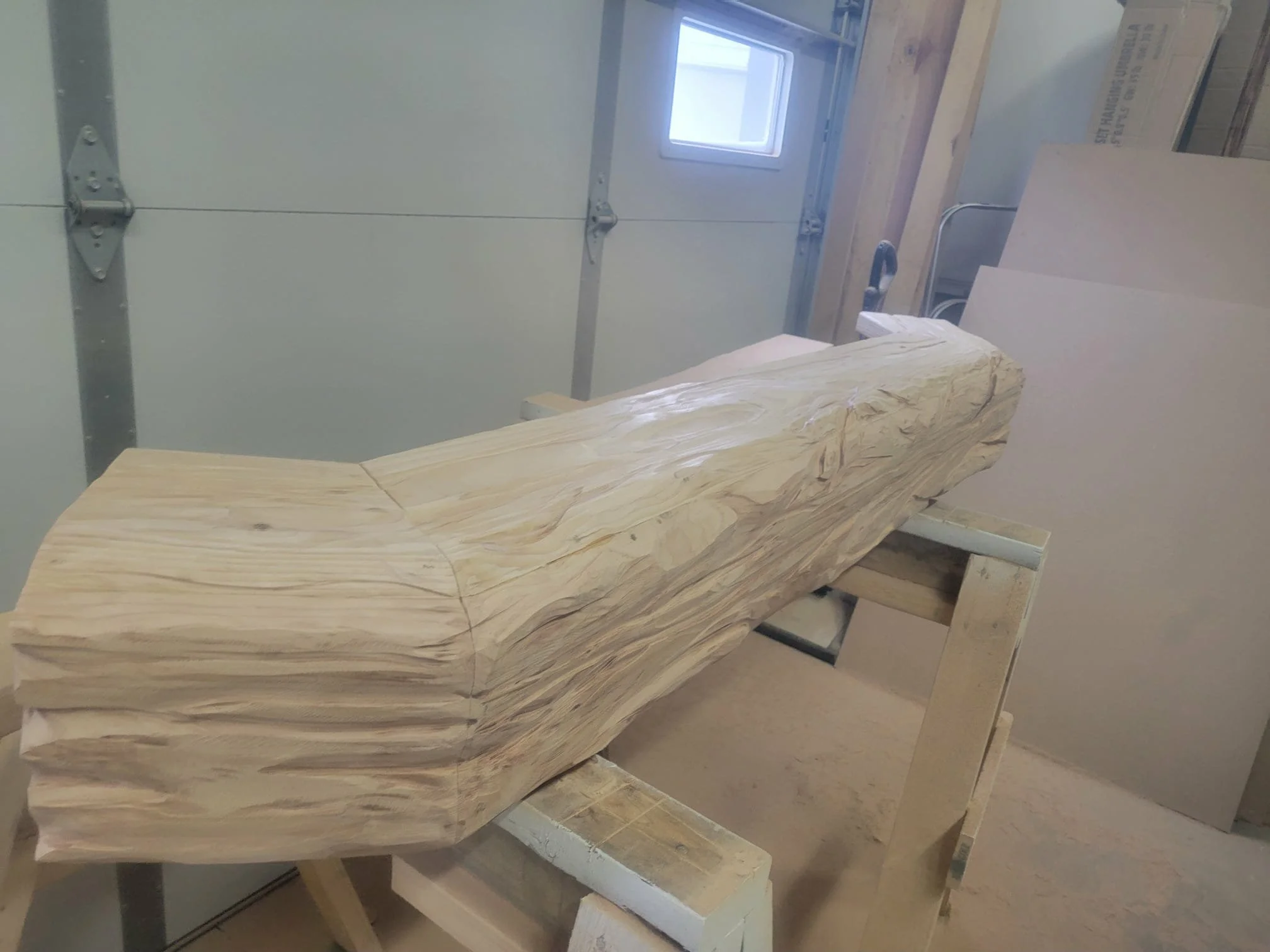 A large piece of light-colored wood with natural grain sits on a wooden workbench in a woodworking shop, with a garage door, window, and various tools and materials in the background.  Custom carved mantle unstained.