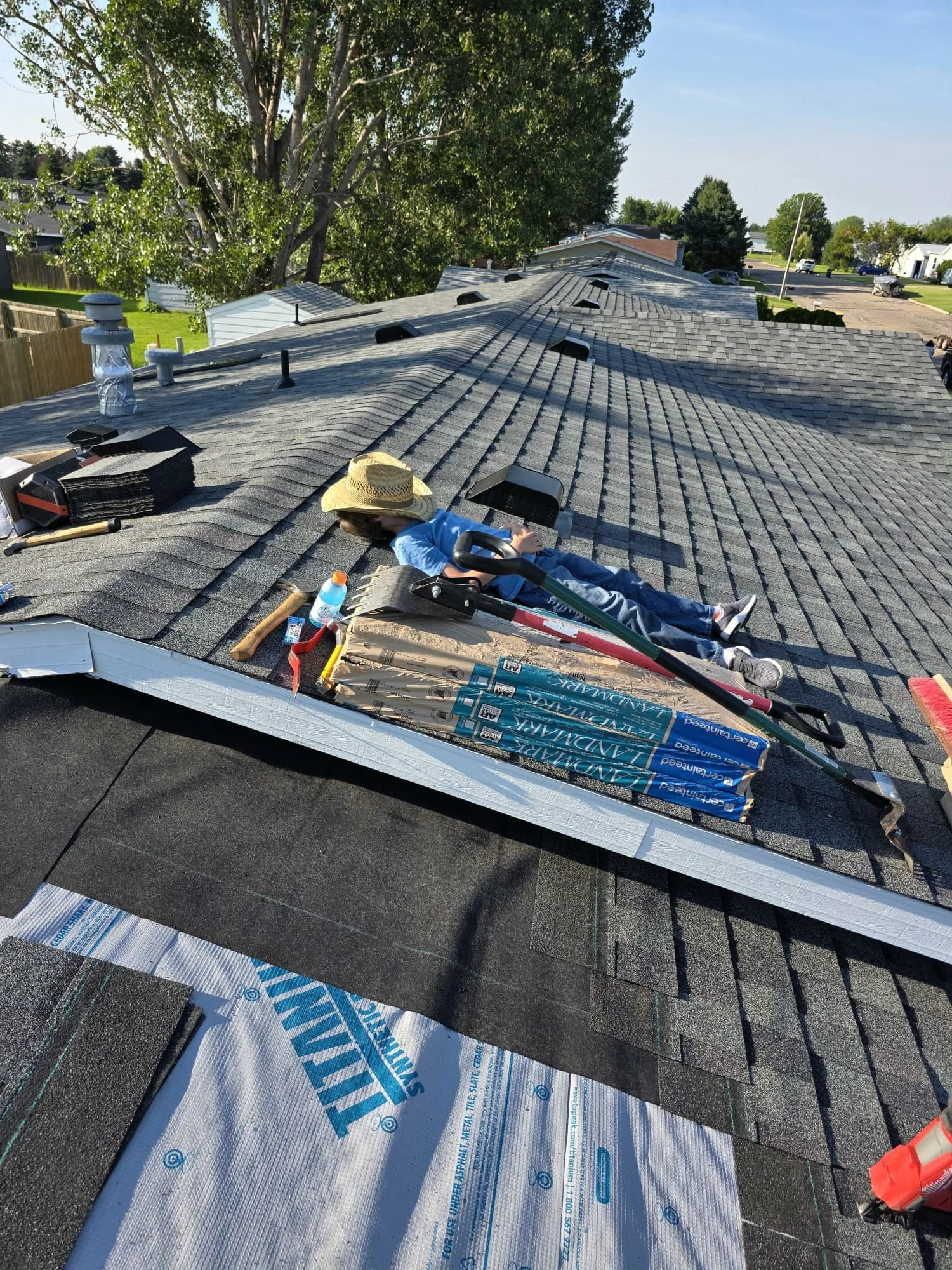 A person lying on a roof installing new shingles with a hat on and tools beside them, with the sky and trees in the background.
