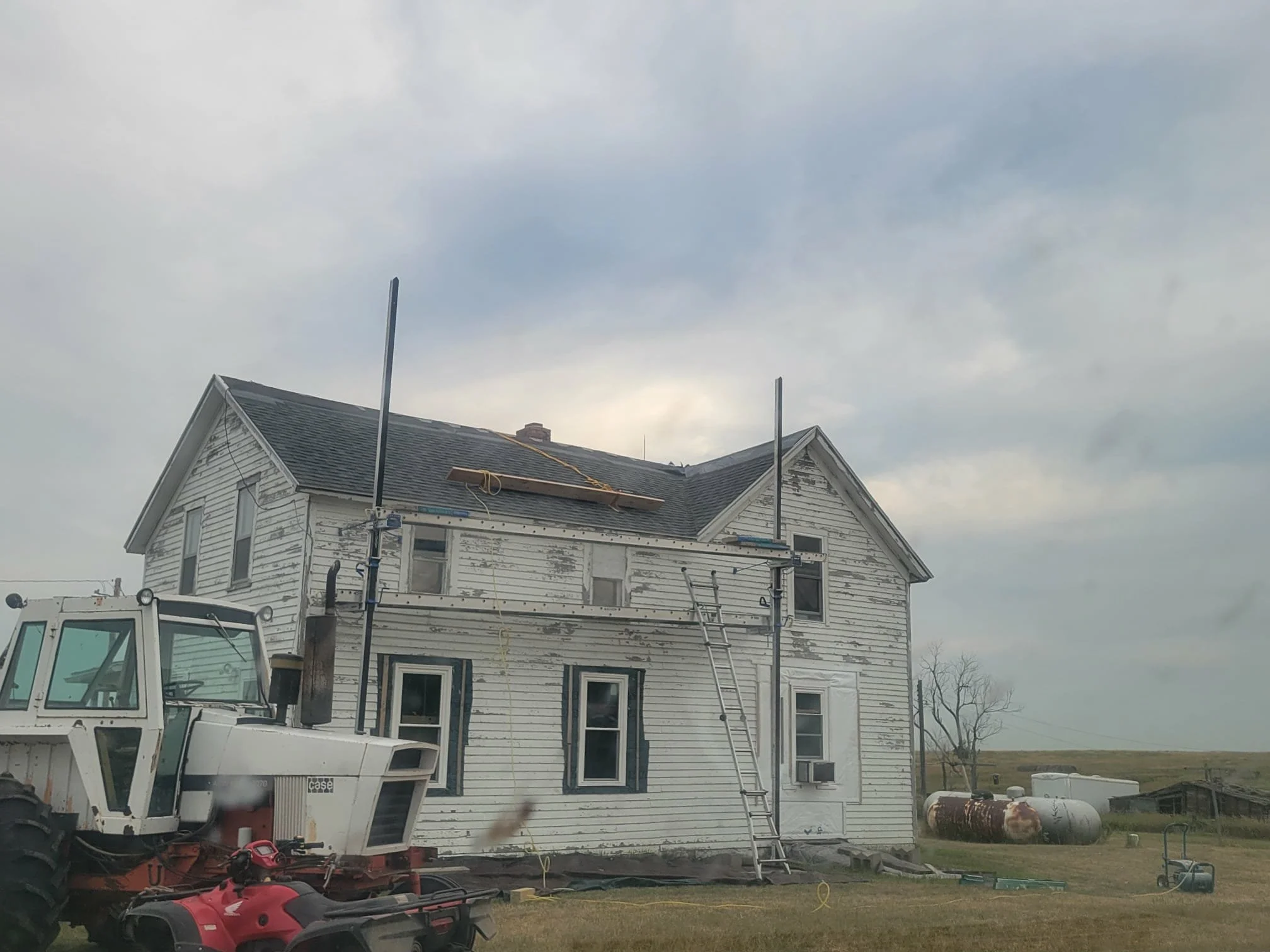A two-story house undergoing repairs with shingles removed from the front and side walls, a ladder leaning against it, and scaffolding with two tall poles in front. There is construction equipment, a tractor, and tanks in the yard on a cloudy day.