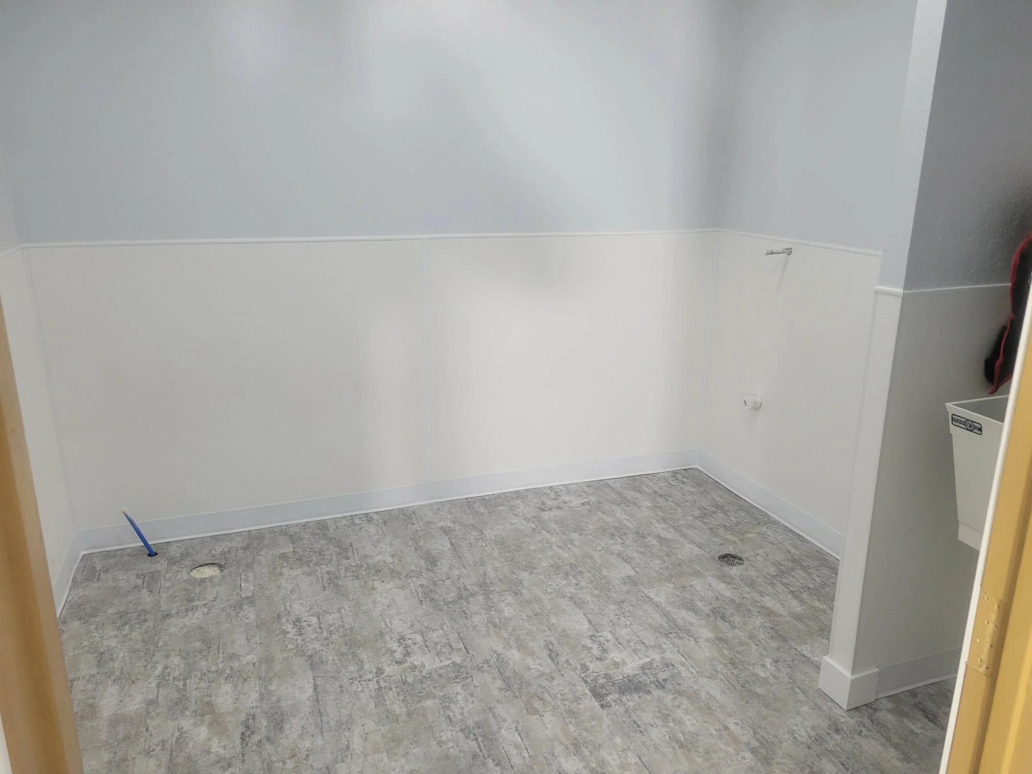 An empty laundry room or utility room with gray flooring, white lower wall panels, and pale blue upper walls. There are plumbing openings on the floor and wall for appliances, and a partial view of a white utility sink or cabinet on the right.