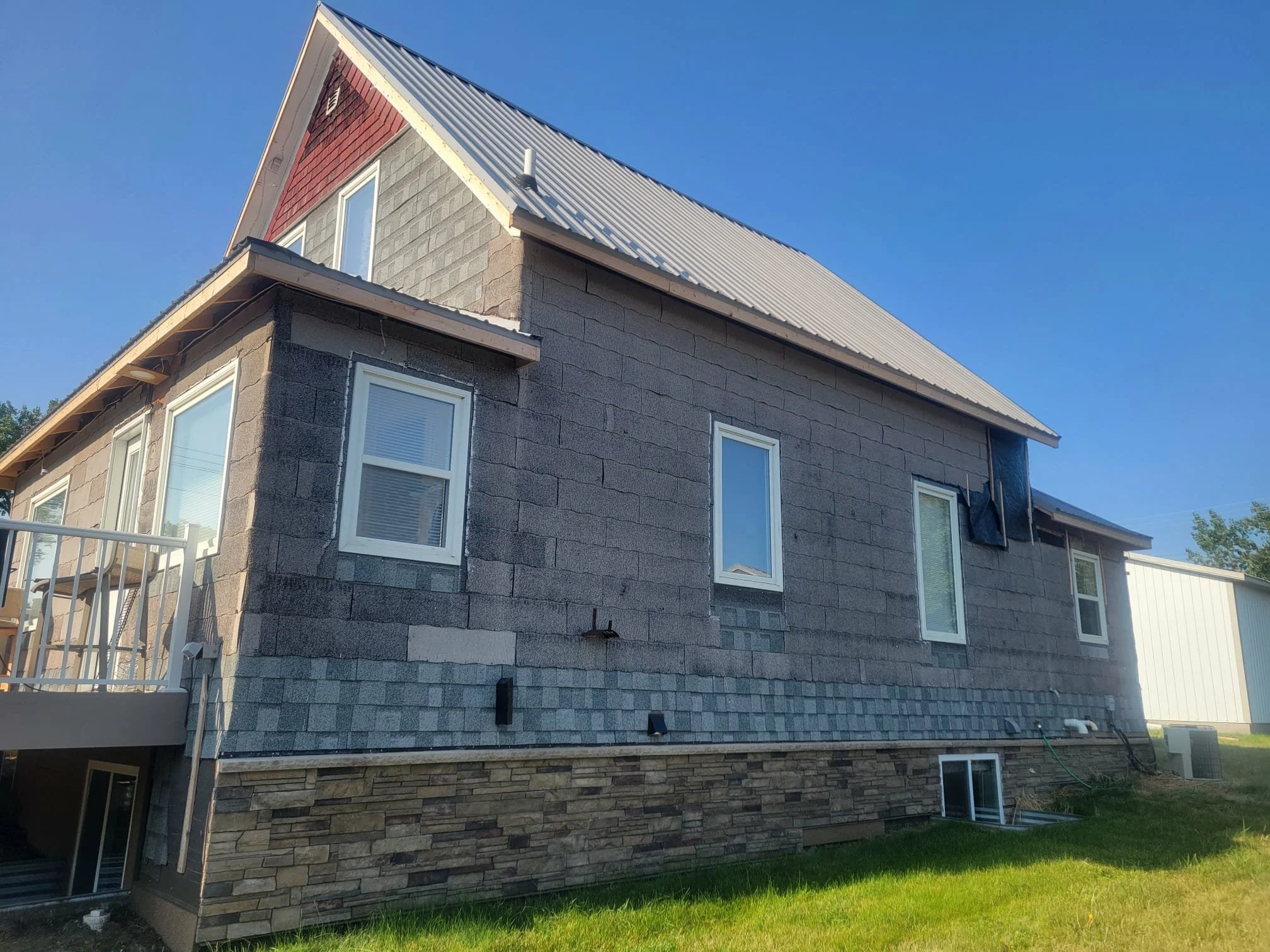 Side view of a two-story house under construction with gray shingle siding, stone veneer on the lower part, white-framed windows, and a metal roof, with a green lawn in the foreground and clear blue sky overhead.