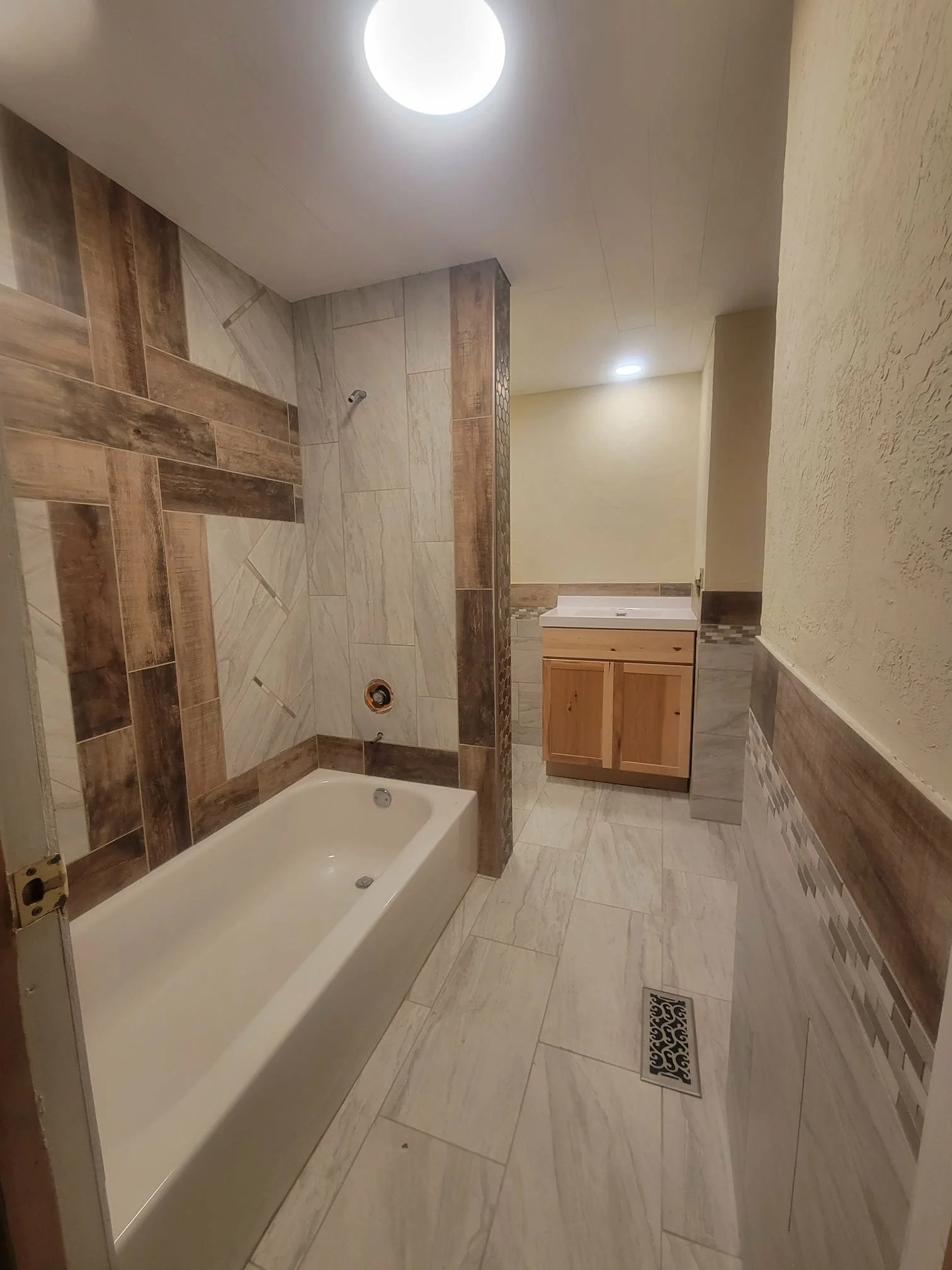 Bathroom with a bathtub, a small vanity with a sink, tiled walls with wood-like and stone-like patterns, and a floor with similar tile patterns.
