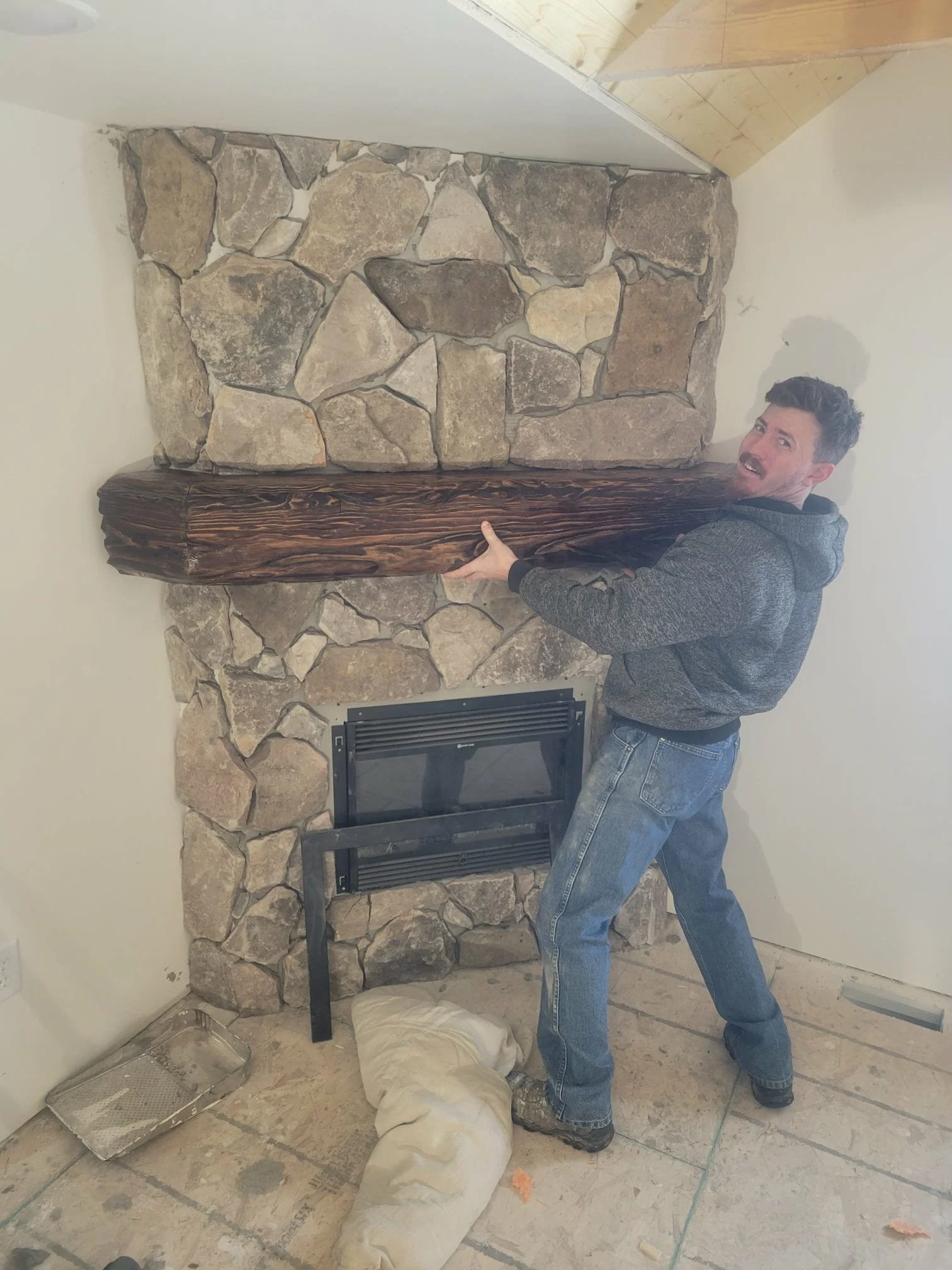 A man in a gray hoodie and jeans is lifting a large custom carved wooden mantle in front of a stone fireplace, with a wall and part of the ceiling visible.