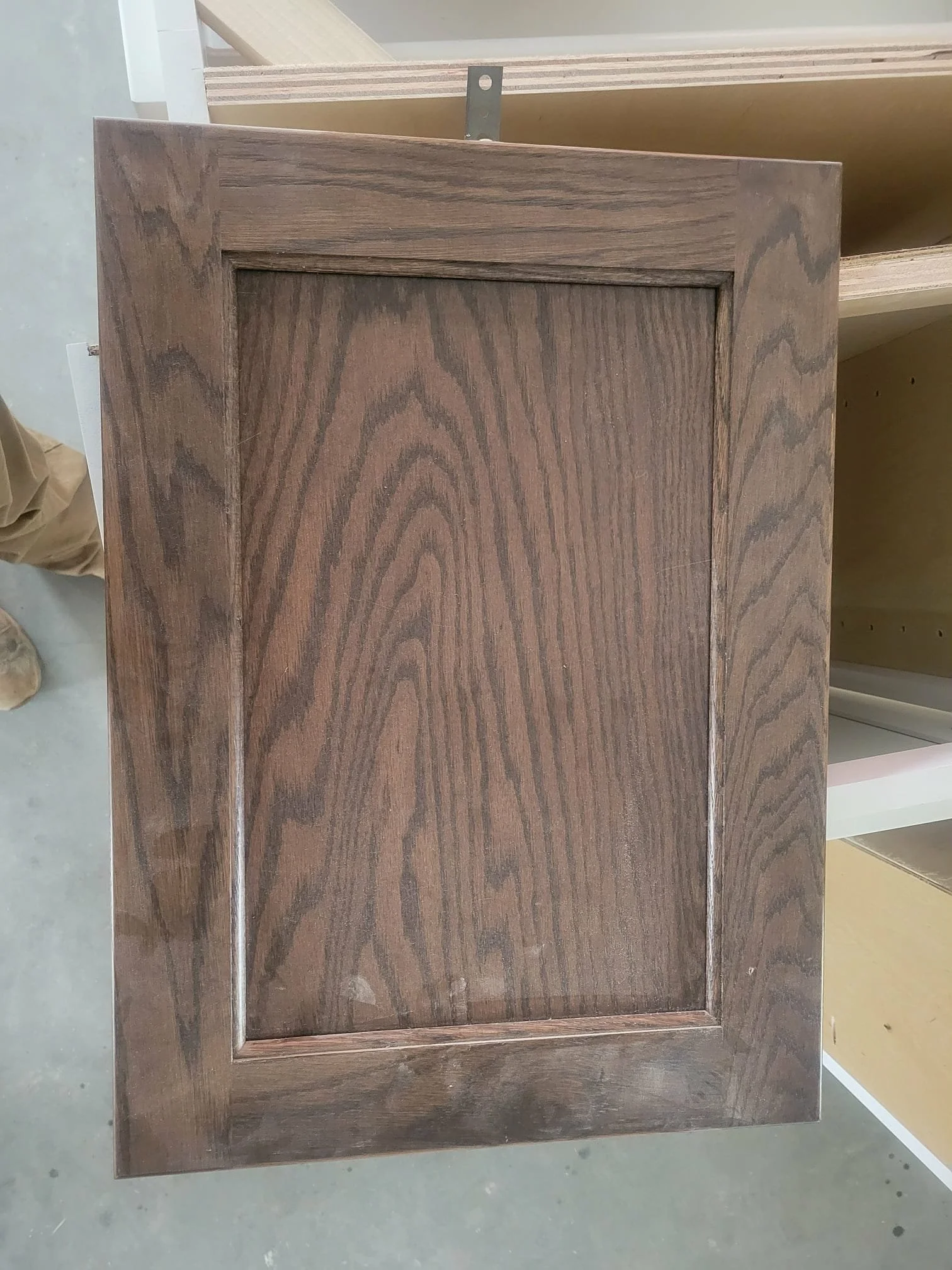 A wooden cabinet door with a raised panel in the center, showing wood grain patterns and a smooth finish, laying flat on a surface.