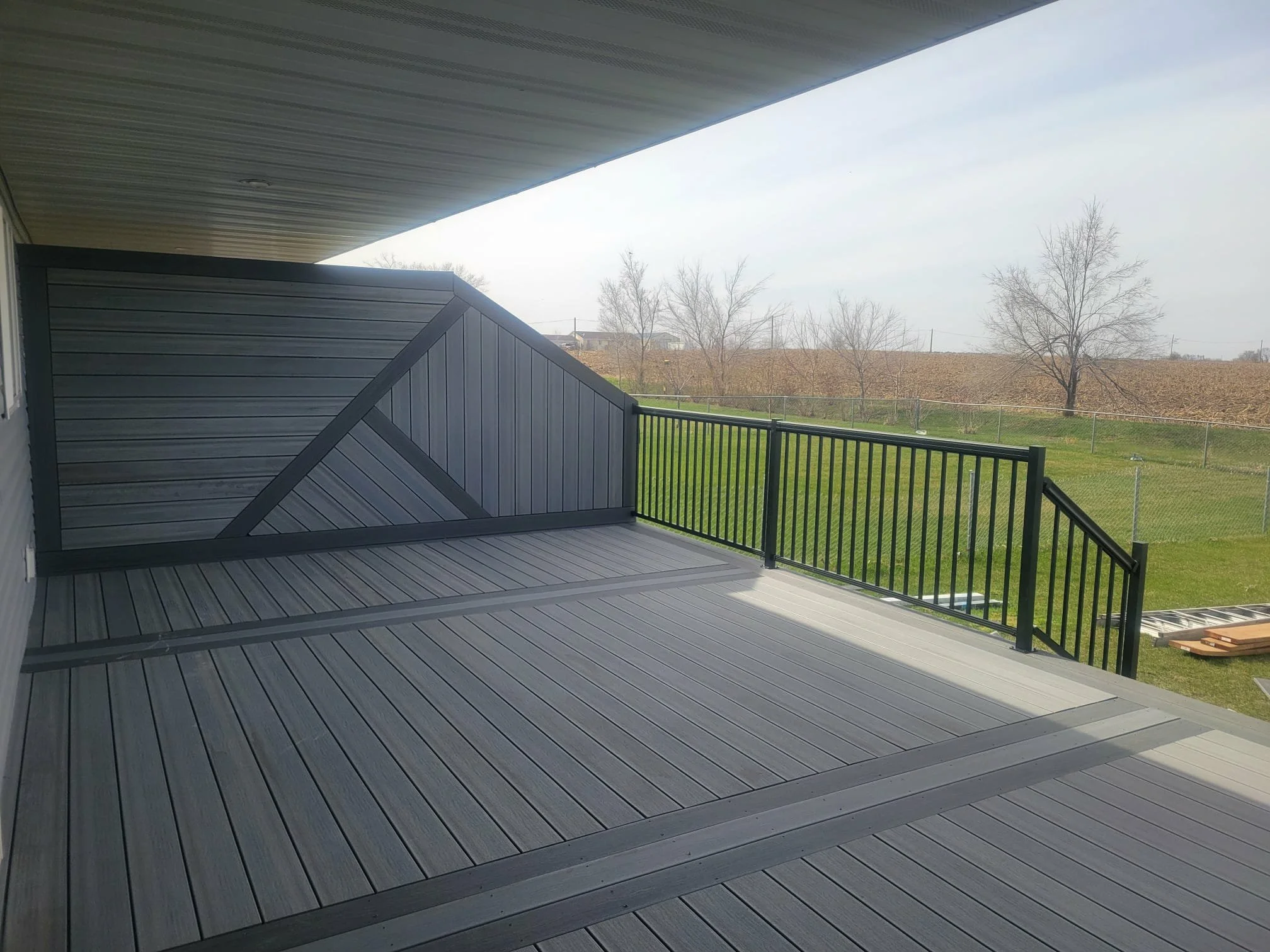 Empty outdoor balcony with a gray wooden floor, black metal railing, and a privacy wall, overlooking a green lawn and leafless trees in the background.