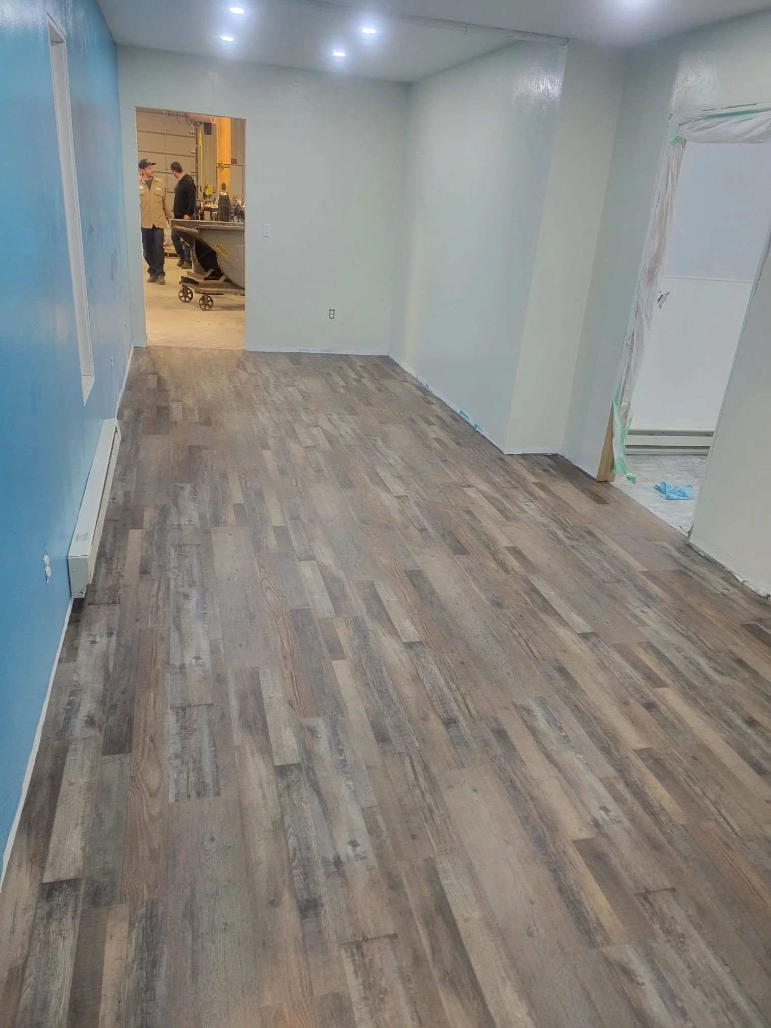 Interior room with newly installed wood-look flooring, white walls, and a partial view of an adjacent room with workers.