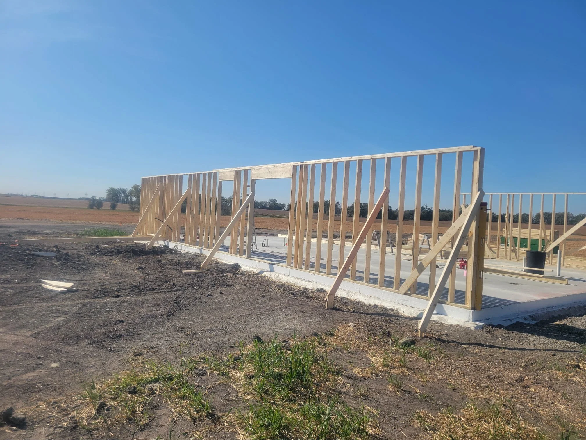 Construction site with wooden framing for a building on a concrete foundation in a rural area under clear blue sky.
