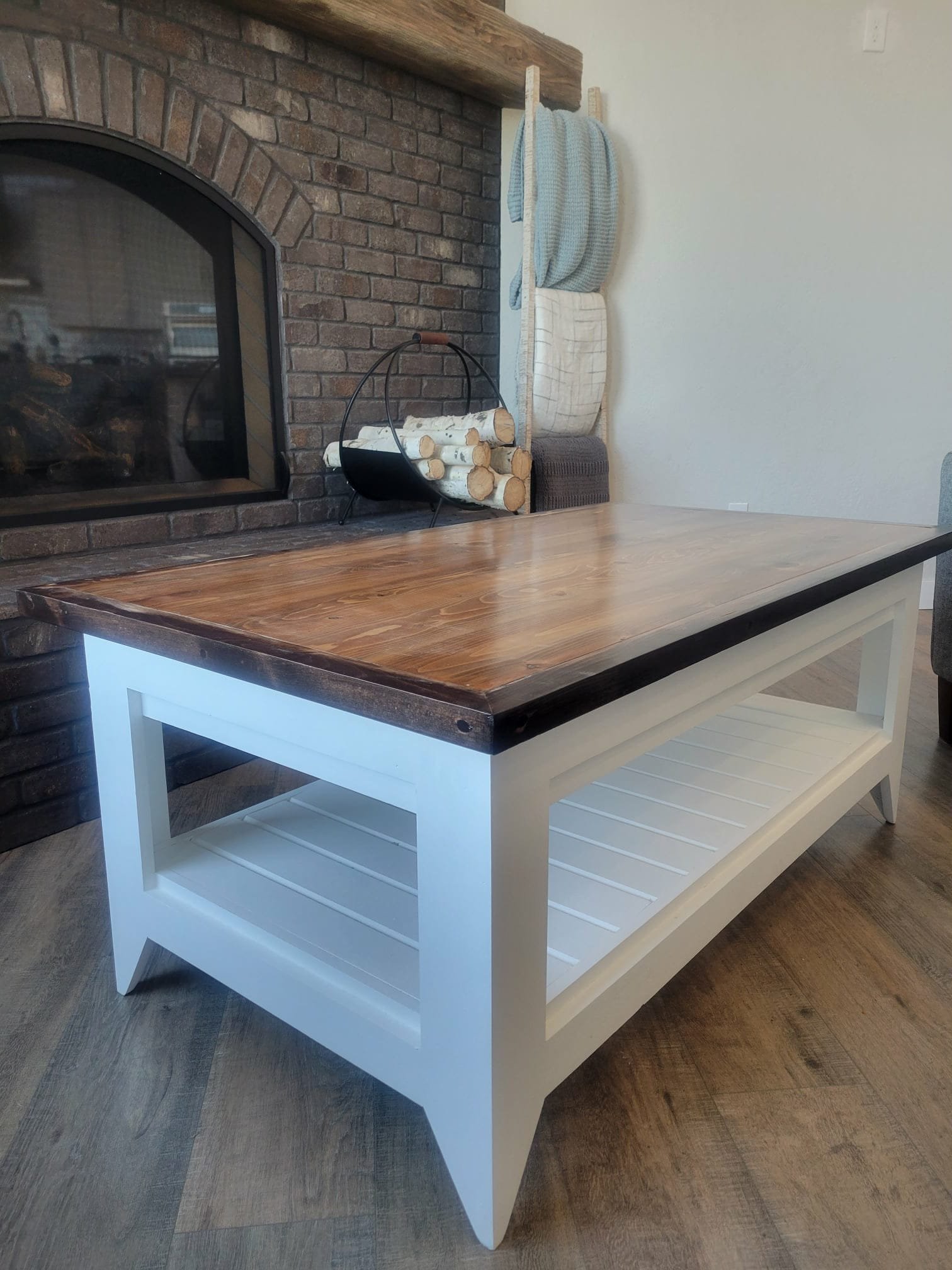 A rectangular wooden coffee table with a dark wood top and white painted frame and lower shelf, situated in front of a brick fireplace.