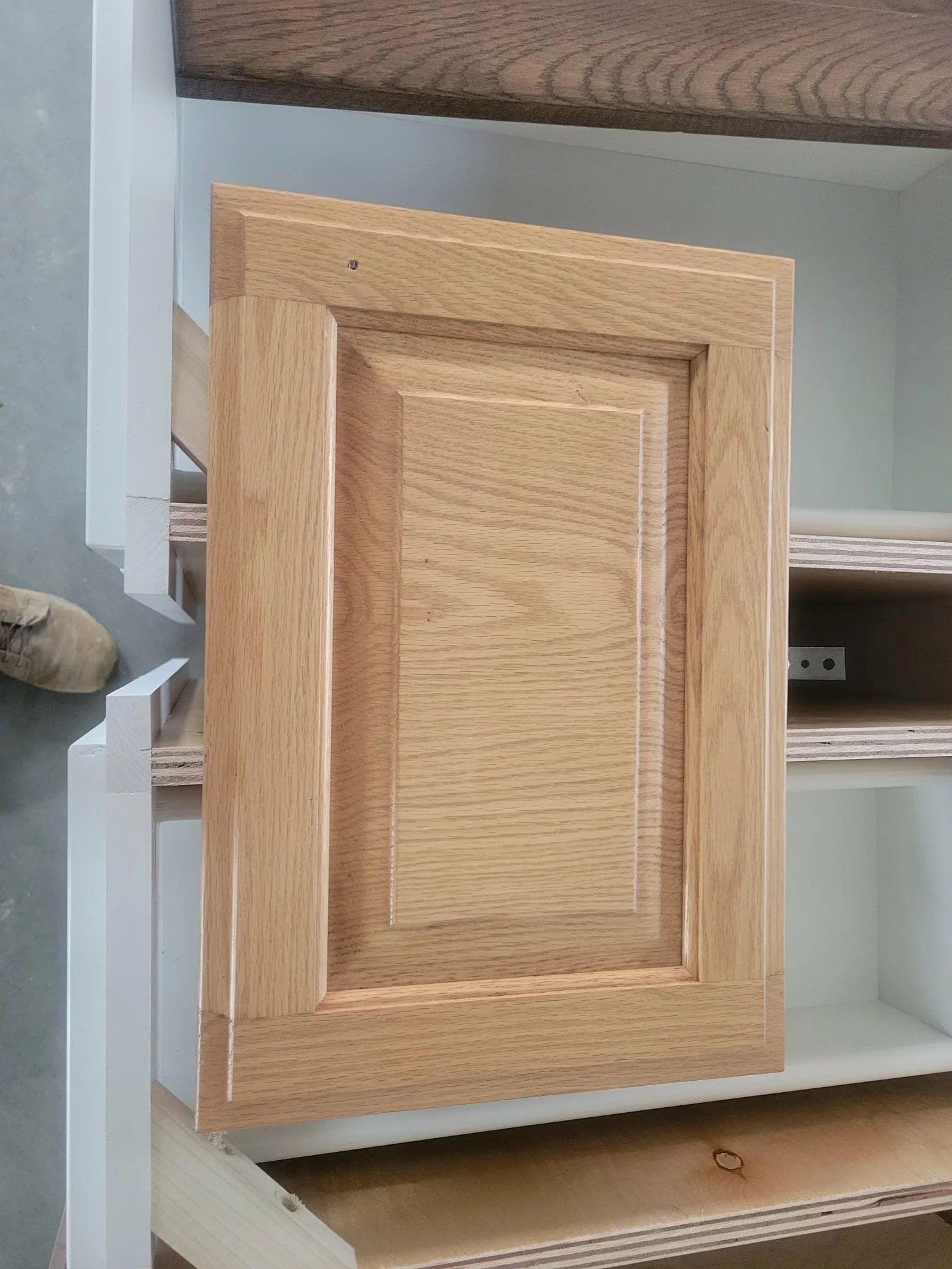 A wooden cabinet door inside a workshop. The door has a raised frame design and is not yet painted or finished.