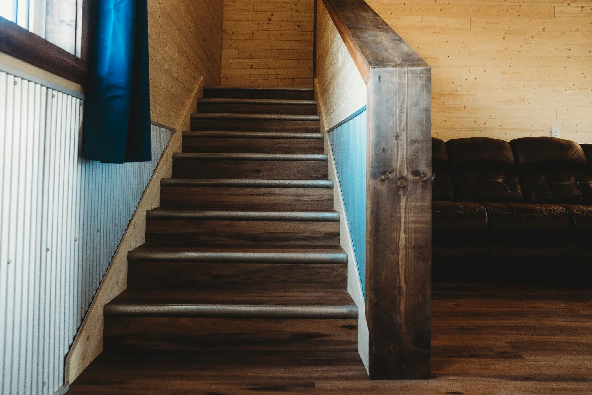 Wooden staircase with dark brown steps and a blue side panel in a cabin interior. To the right, a black leather sofa against wooden paneled walls. A window with a dark blue curtain on the left.