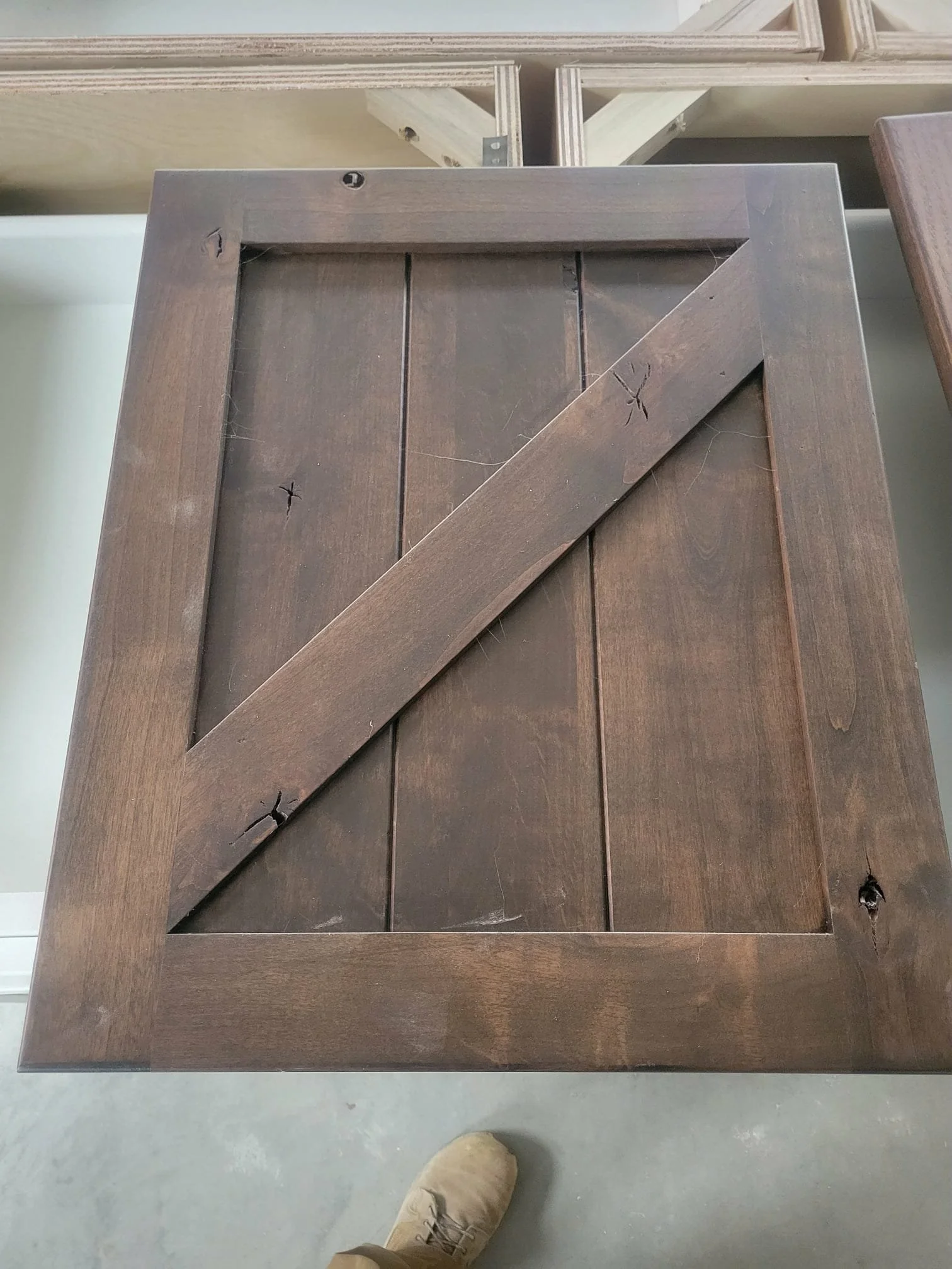 A dark brown wooden cabinet door with a diagonal support strip across the center, three vertical planks behind it, and visible knots and grain, seen from above in a workshop or store.