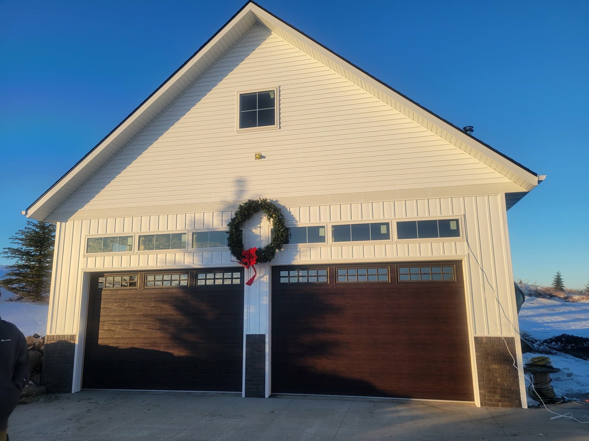 A white garage house with a Christmas wreath hanging above the garage doors, decorated with a red bow, in a snowy landscape during daytime.