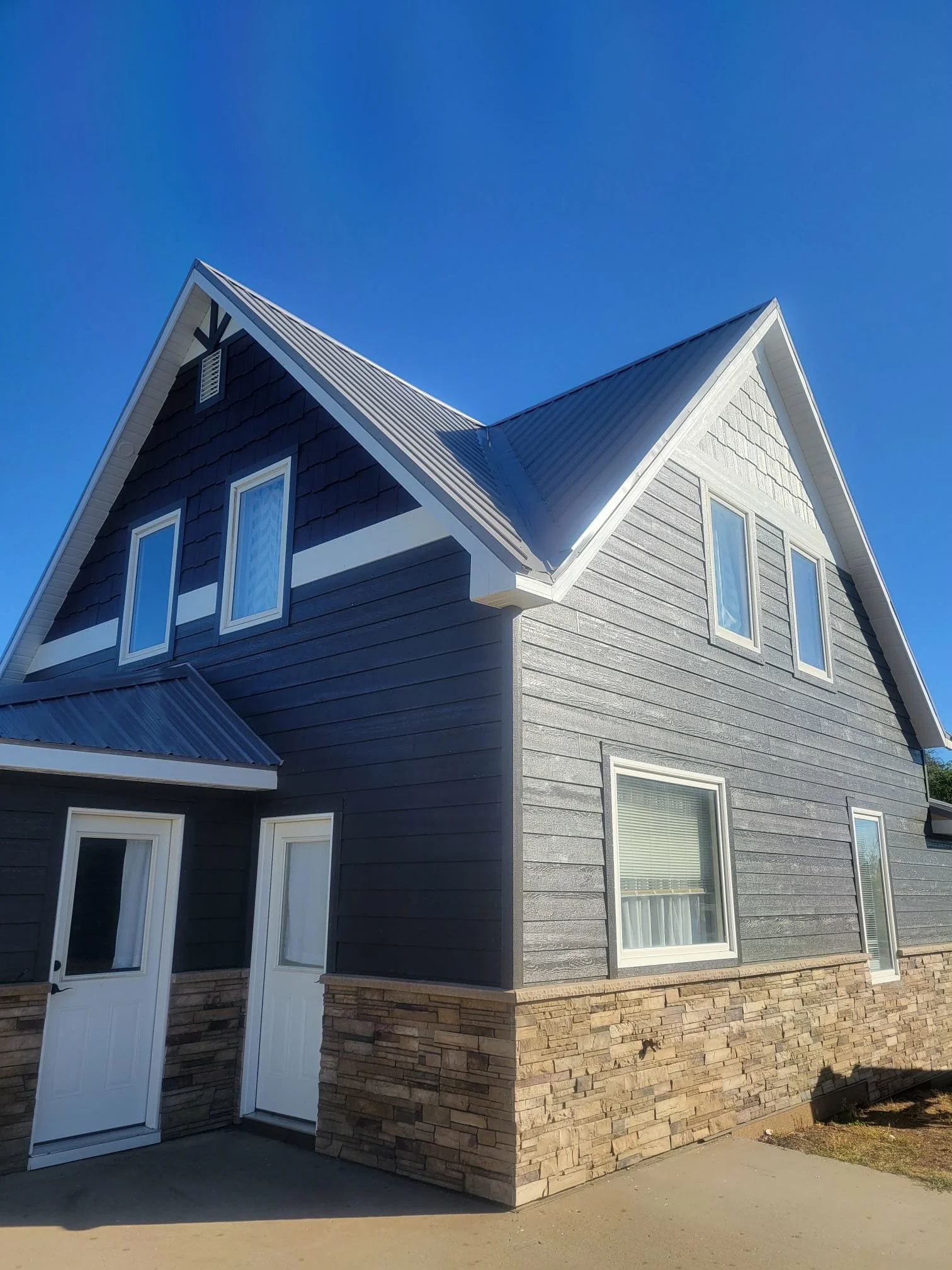 A two-story house with dark horizontal siding, a stone foundation, metal roofing, and multiple windows under a clear blue sky.