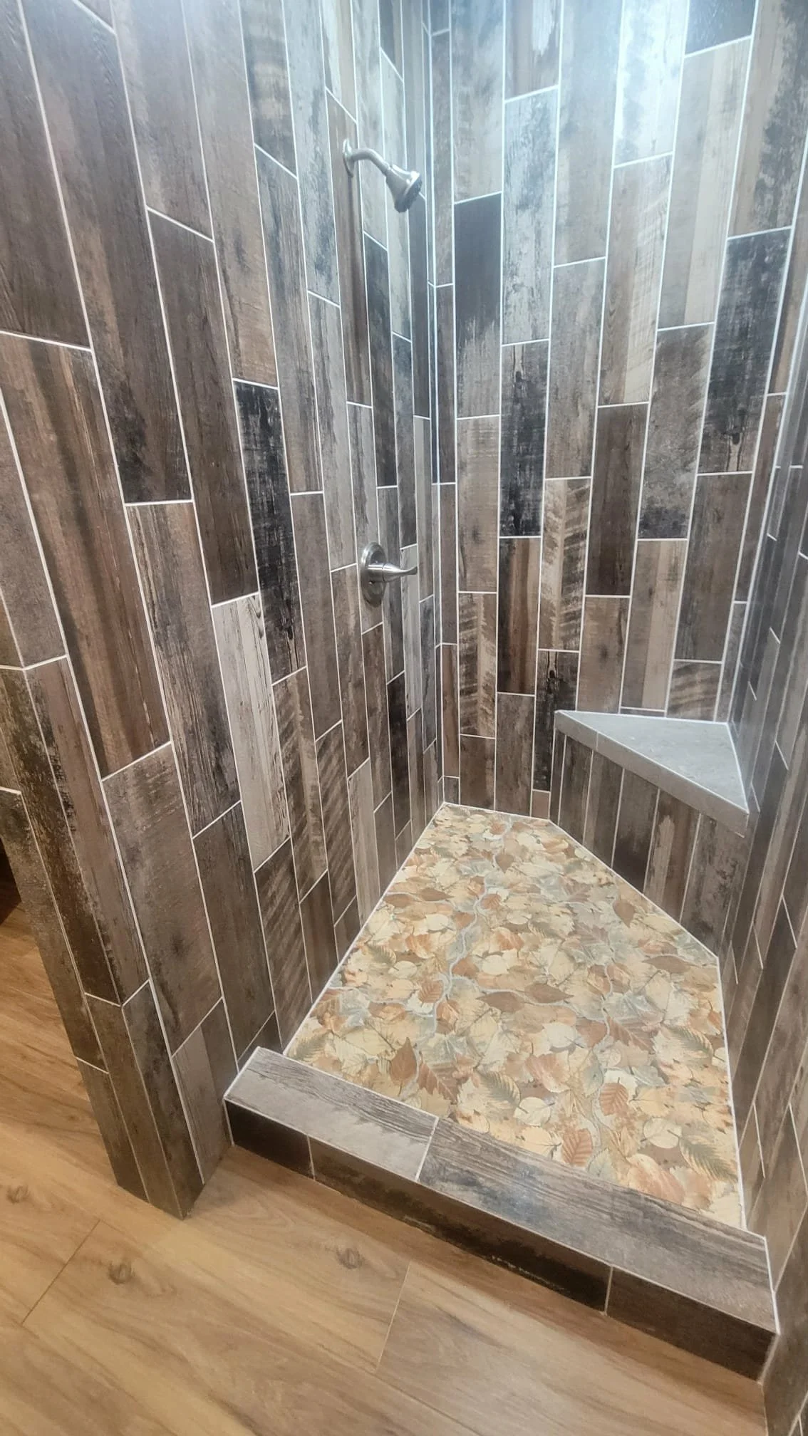 A walk-in shower with brown and gray patterned tile walls, a built-in corner seat with a gray tiled surface, and a floor with a leaf pattern design in earthy tones.