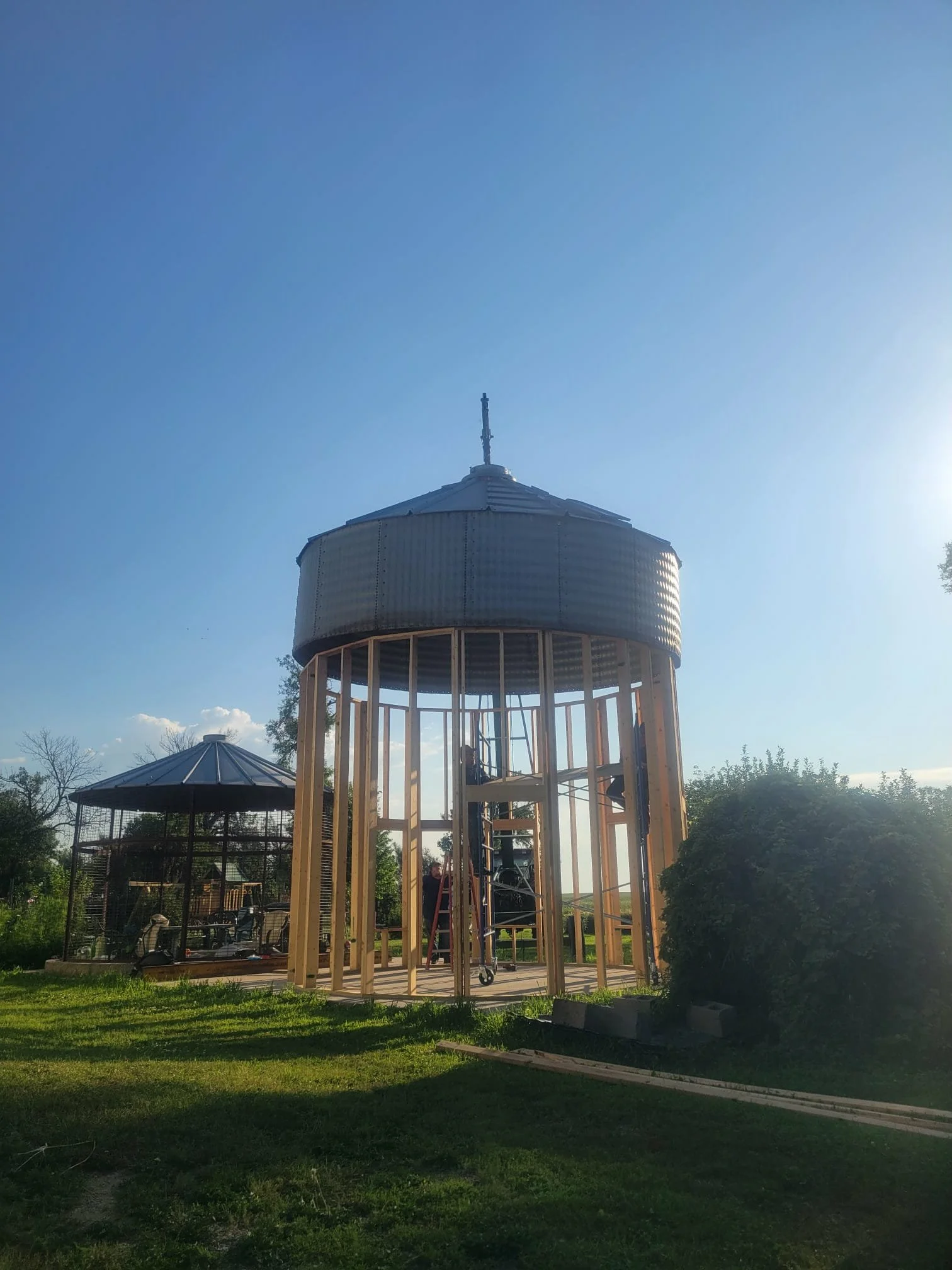 A partially constructed wooden and metal gazebo with a round upper section and a cone-shaped roof in a grassy outdoor area.