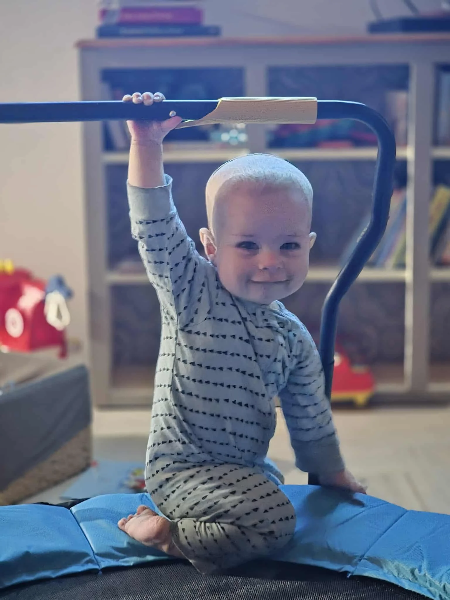 A young child with short blonde hair balancing on a trampoline pad, gripping a handlebar for support, in a living room with a bookshelf and toys in the background.