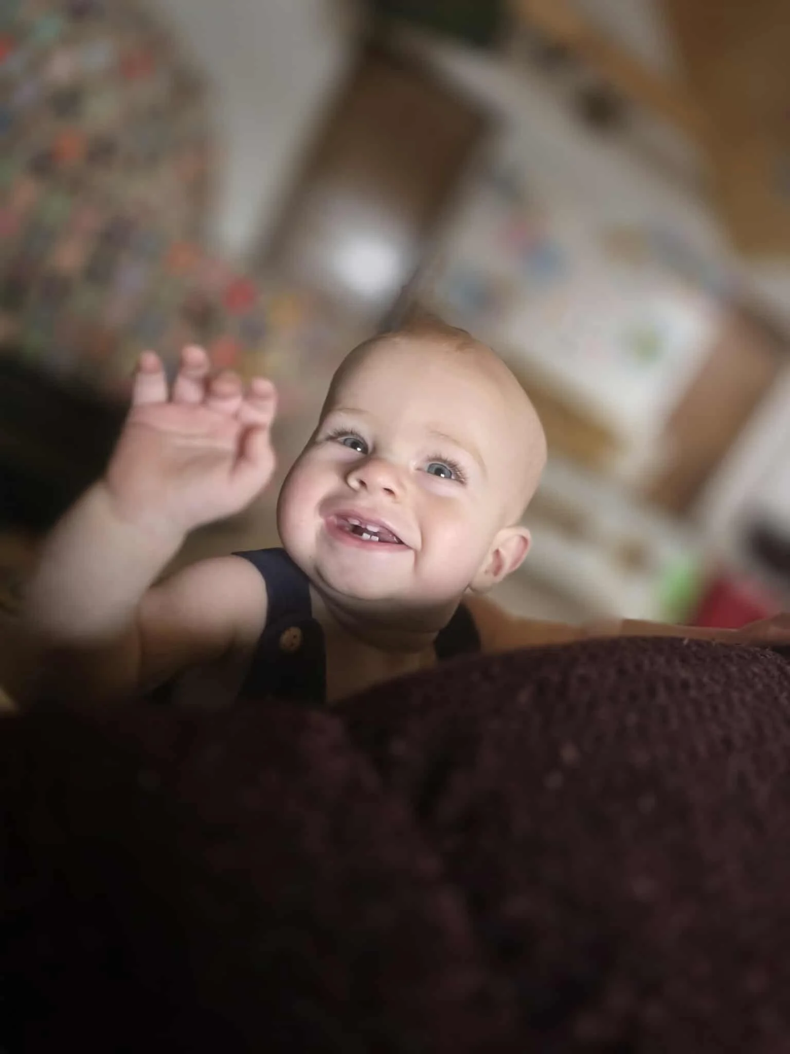 A smiling baby with missing front teeth, reaching up with one hand, indoors with blurry furniture and a wall in the background.