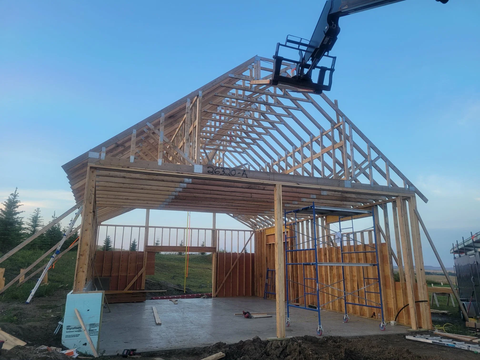 A house under construction with a wooden frame, scaffolding, and a lift. The roof structure is in progress with exposed trusses and a concrete slab is visible on the floor. The background shows a clear sky and distant trees.