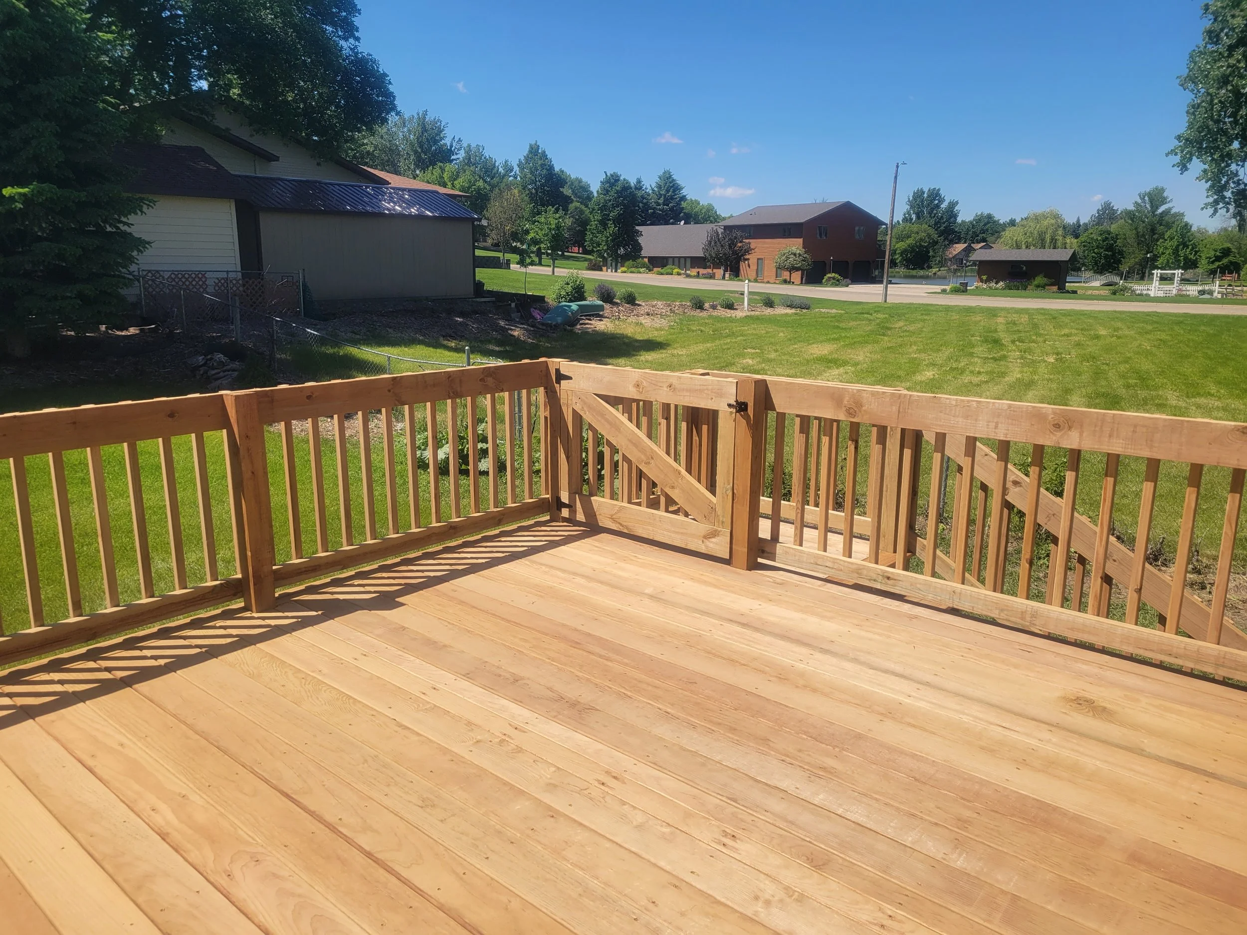 View of a wooden deck with a grass lawn, houses, trees, and a road in the distance on a sunny day.