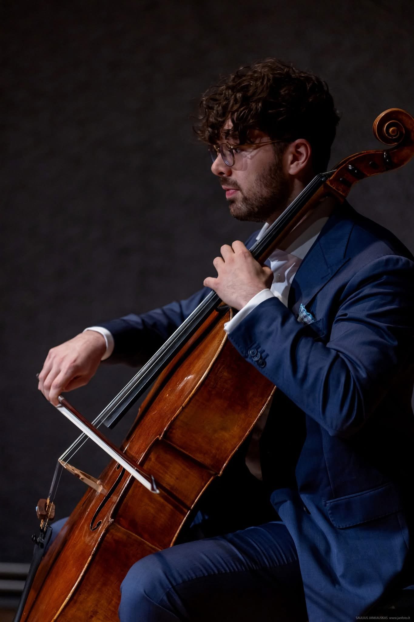 A man with curly hair, glasses, and a beard dressed in a blue suit playing a cello against a dark background.
