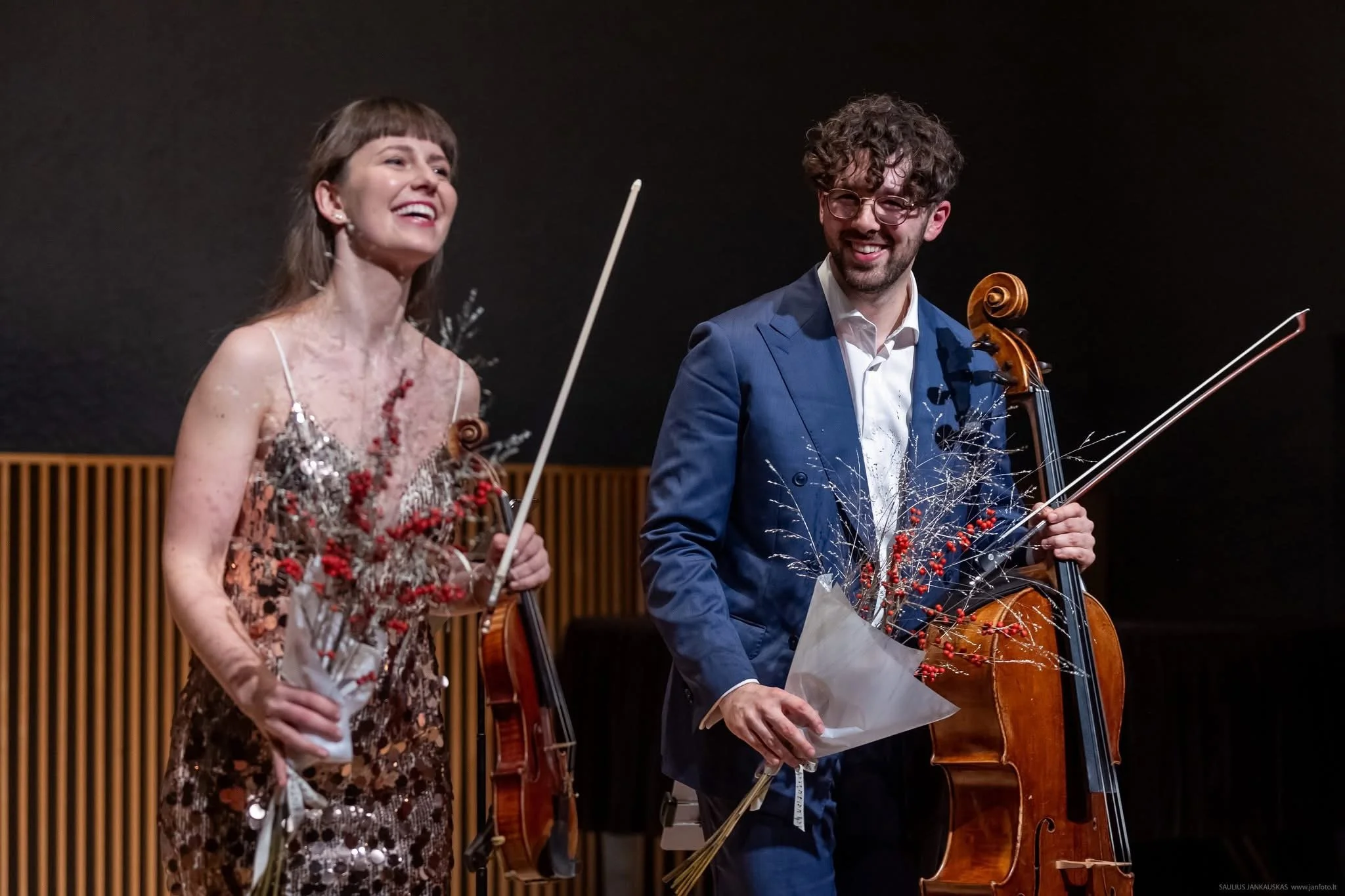 A man and woman holding violins, flowers, and smiling after a performance, standing in front of a black background.
